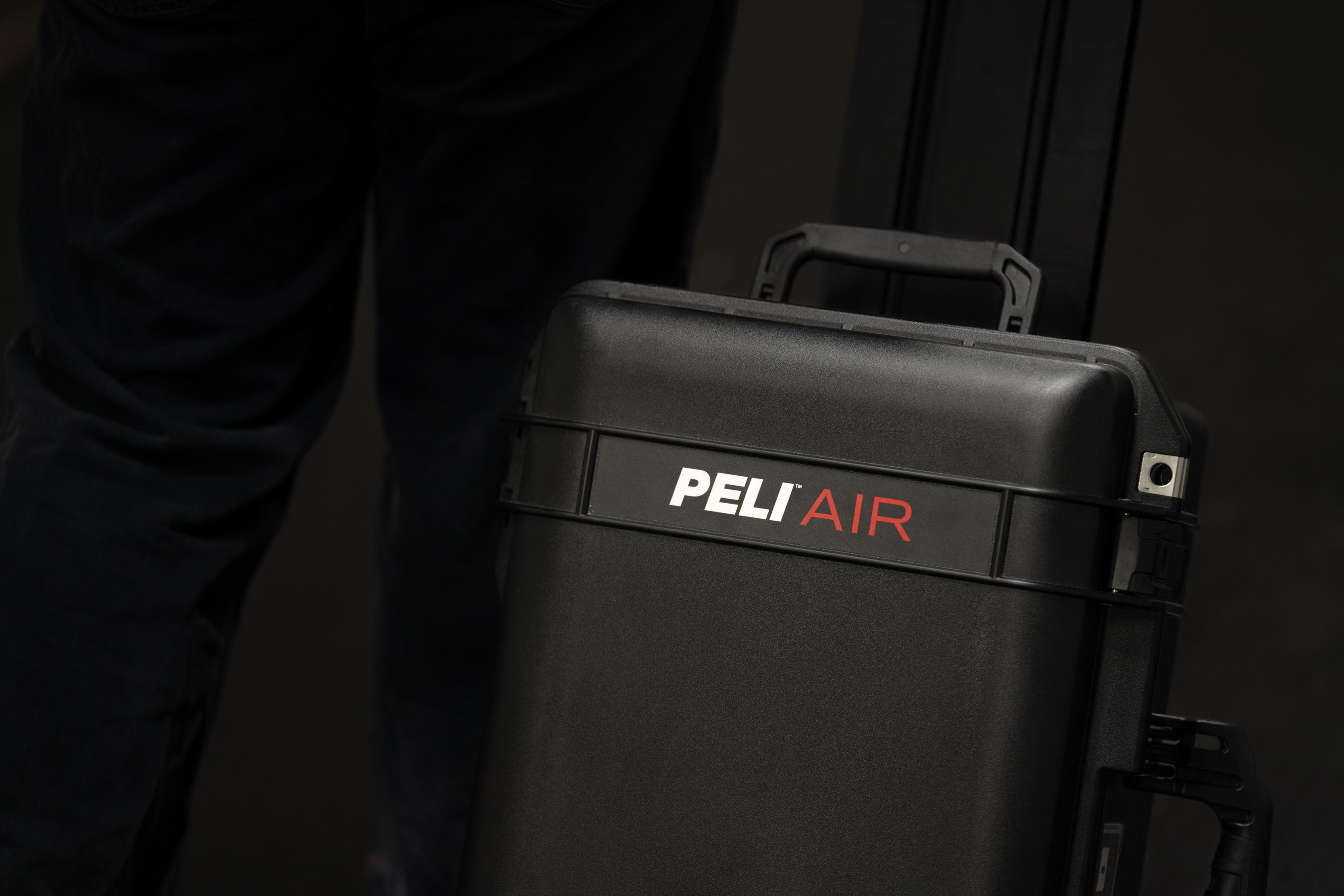 A model, wearing black jeans, carries the Peli 1535 Air hard case, against a black background, displaying the prominent Peli Air logo
