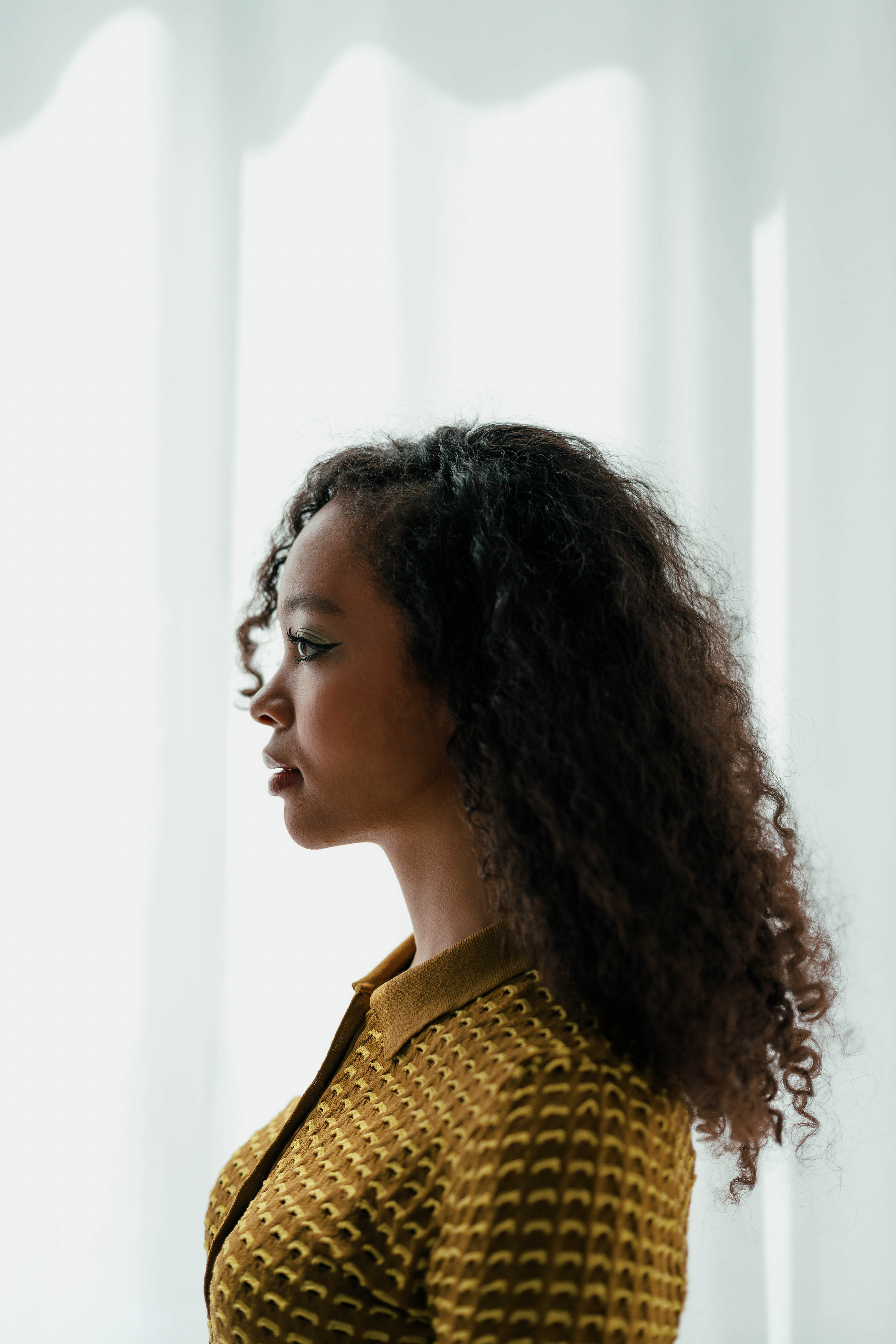 A softly-lit profile shot of a model against a bright window with a white curtain. Shot by Alina Rudya on a Nikon Z6 III.