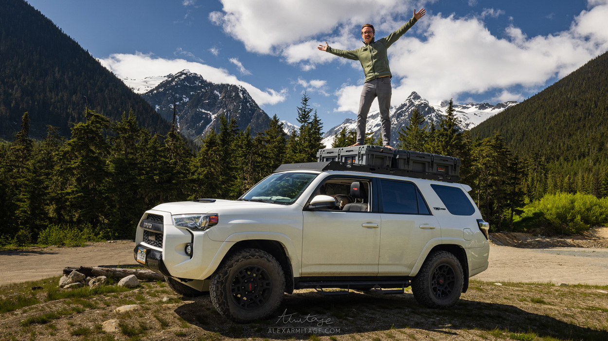 A person stands with their arms up while standing on a vehicle in the mountains.