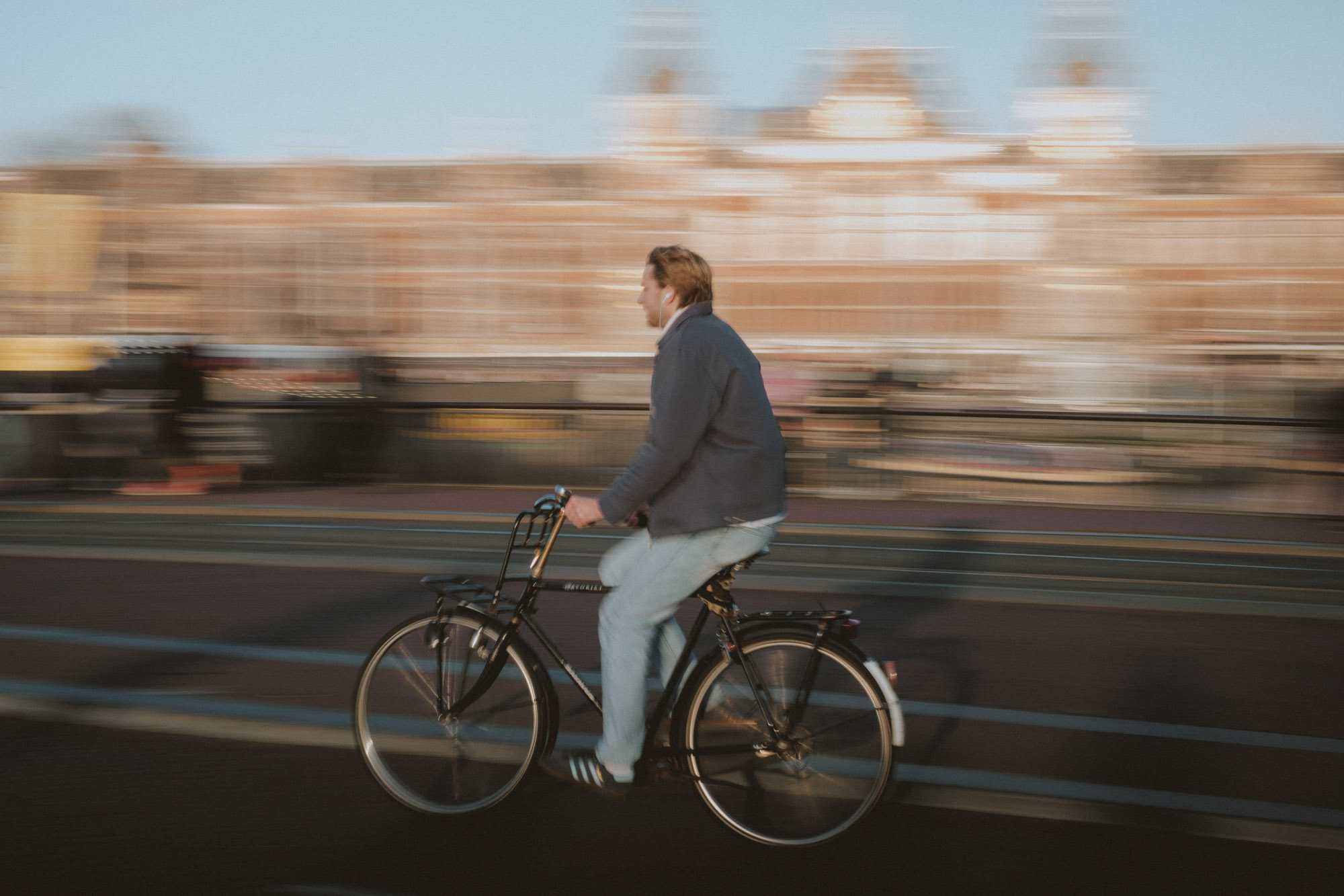 Man cycling across an Amsterdam bridge captured with motion blur, with historic Dutch architecture in the background during warm evening light.