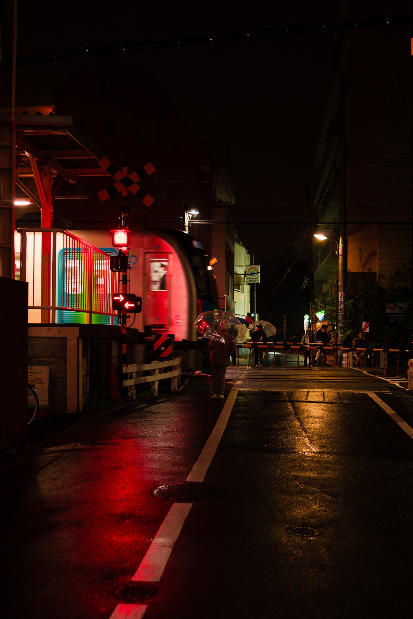 An atmospheric scene of a train crossing in Japan at night, illuminated by red lights and reflections on the wet pavement, with pedestrians holding umbrellas in the rain. Photographed by Ruby Cruden with a Sony DSC-RX1 RM II.