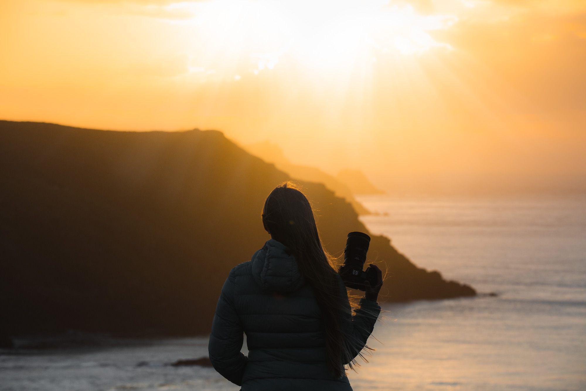 A silhouette of a person holding a camera, with bright sunlight, a coastline and a cliff in the background.