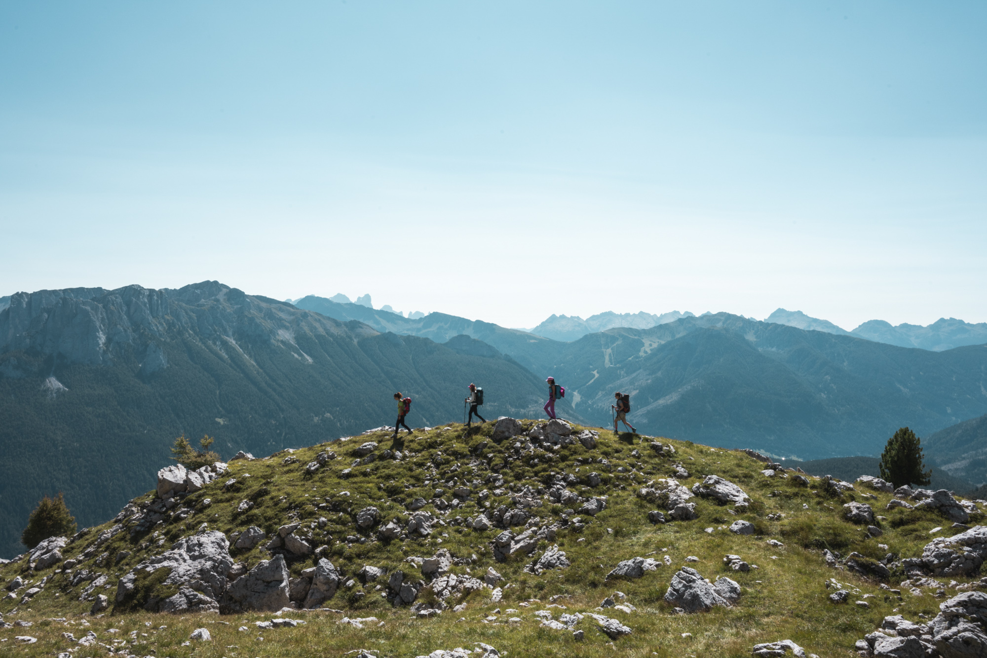 Vier wandernde Personen mit Rucksäcken, die der Reihe nach einen Felskamm mit Alpenpanorama im Hintergrund überqueren.