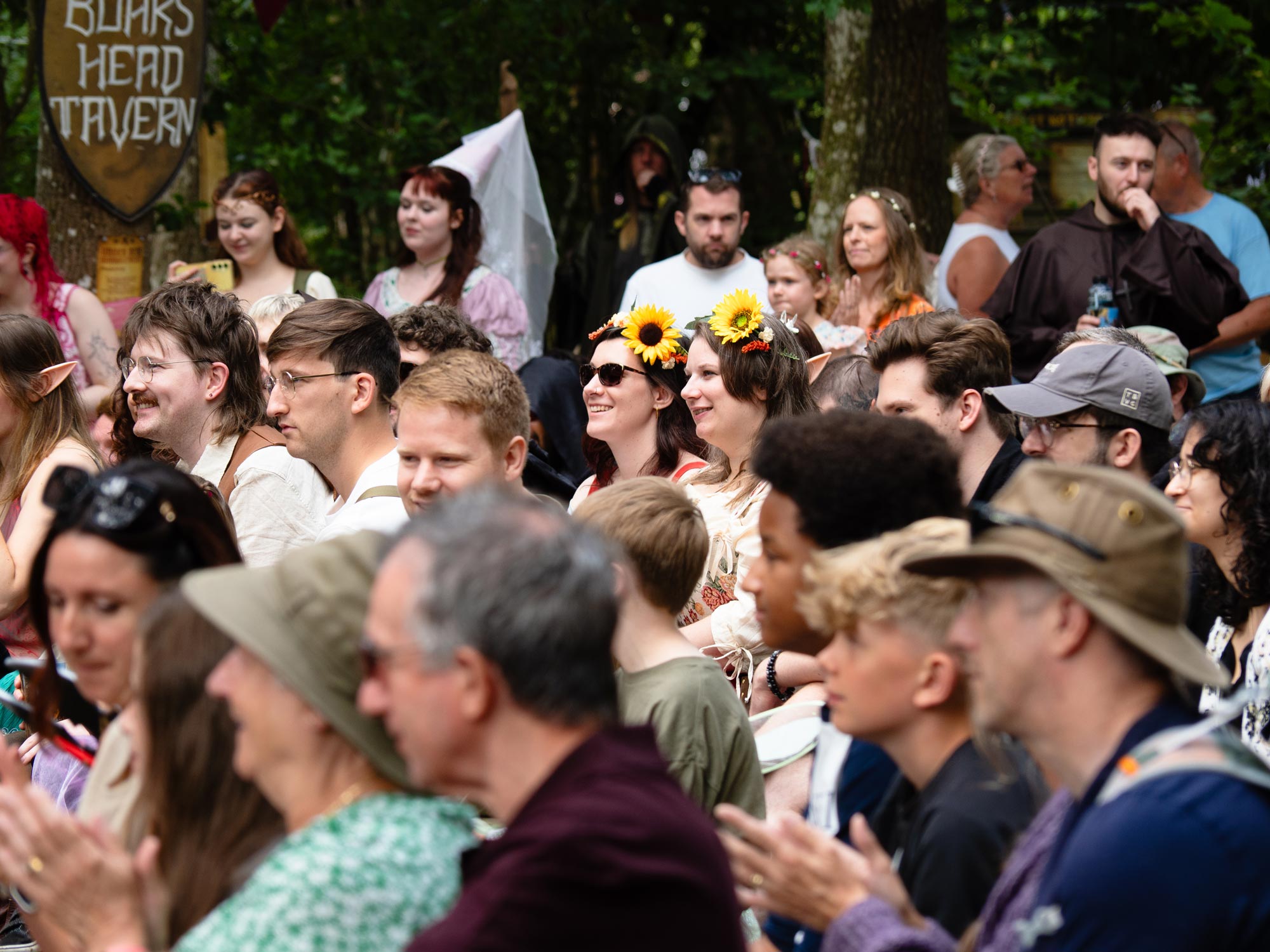 A crowd watches a competition at a medieval-themed event. Two women sit centre frame wearing sunflower headbands.