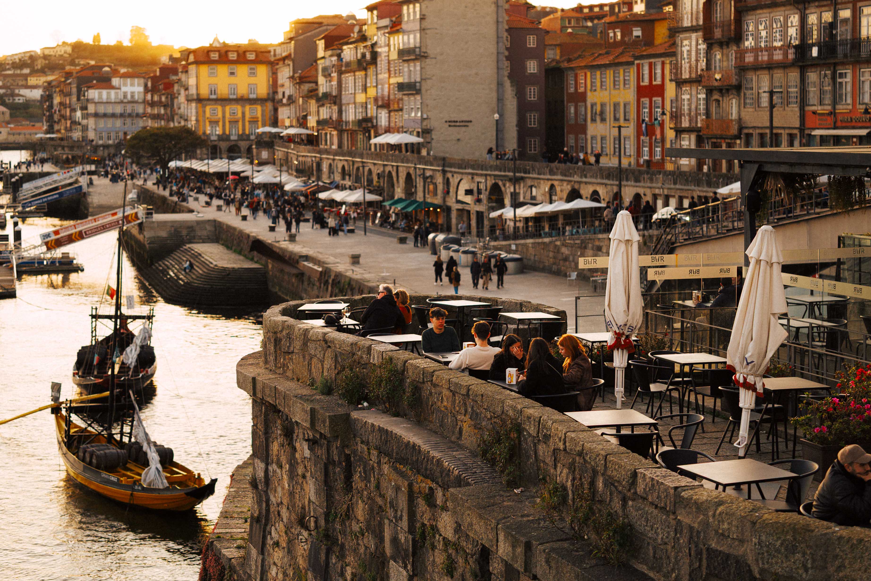 People sitting at outdoor café tables along the Ribeira waterfront at sunset, overlooking the Douro River with traditional boats moored below and colourful buildings glowing in golden light.
