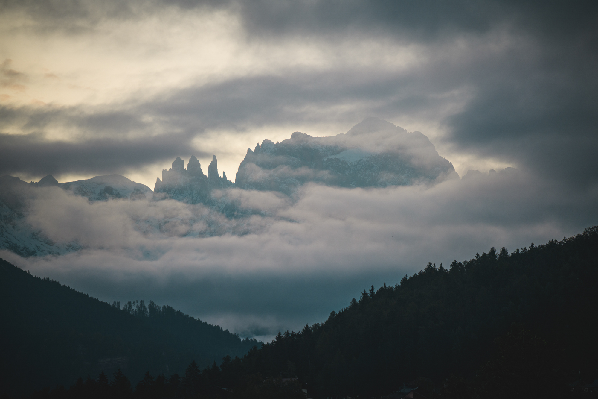 Verschneite Dolomitengipfel, die in einer Winterlandschaft mit Nadelwald im Vordergrund aus einem Wolkenmeer emporragen.