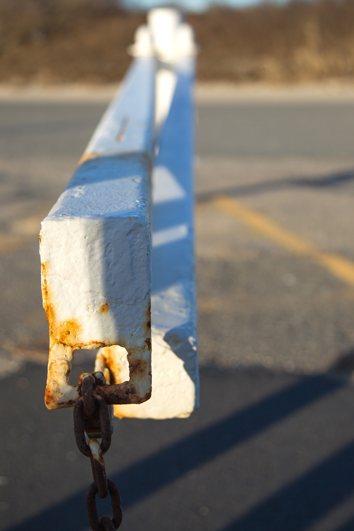 A white gate with a rust chain on the end. There are trees in the background and yellow markings on the road below. Shot by Kim Fuller.