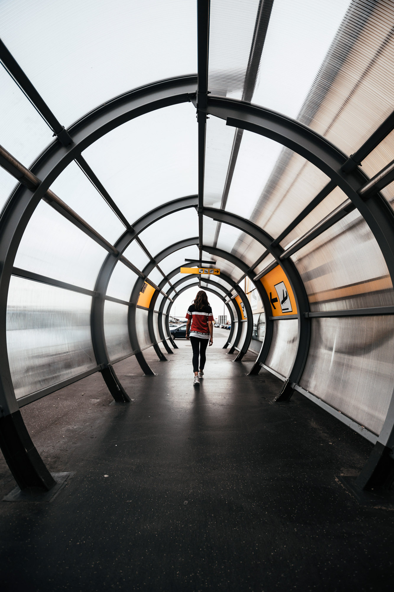 Symmetrischer, tunnelartiger Fußgängerdurchgang am Flughafen Wien mit runden Metallbögen und Fluchtlinien bis zum Ausgang. Darin eine Person, die Richtung Ausgang läuft. Aufgenommen von Dhan Limwattana.