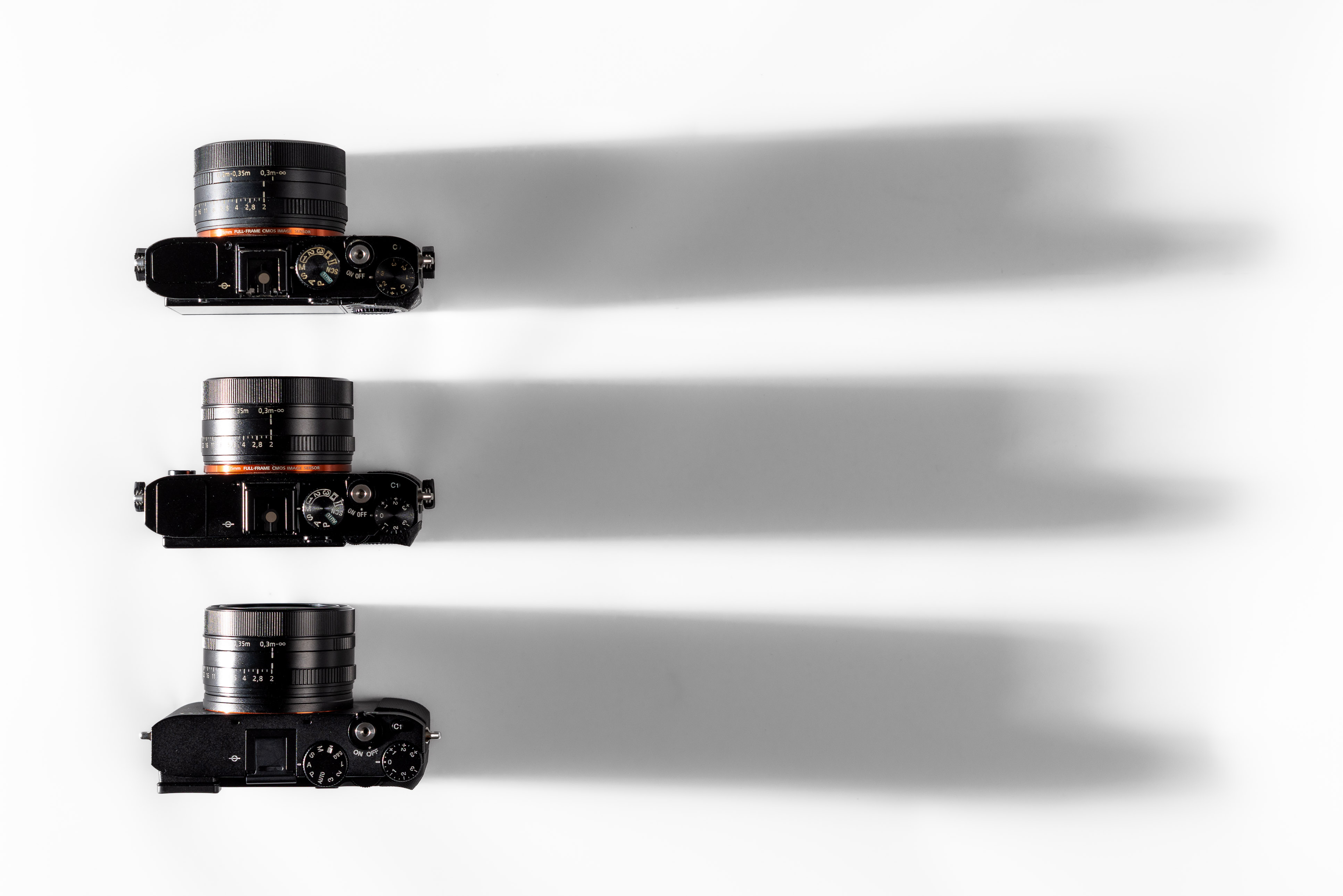 Drei Kameras der Sony RX1R-Serie, die vertikal auf weißem Hintergrund angeordnet sind und lange Schatten nach rechts werfen. Auf diesem Foto sind die Unterschiede bei Gehäusegröße und Anordnung der Bedienelemente zu sehen.