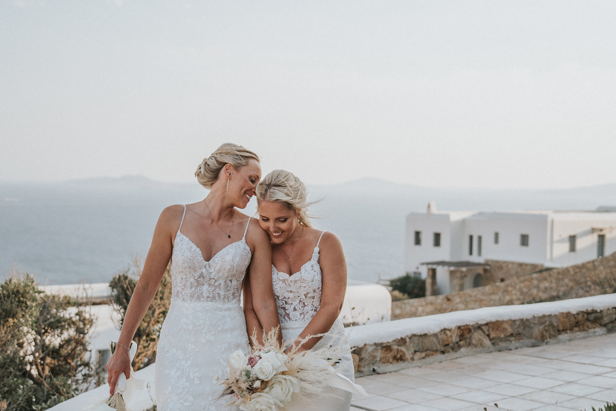 Newlyweds in white wedding dresses, carrying a bouquet of flowers, holding hands and smiling in front of a scenic view of mountains and villas. Shot by Steph Large