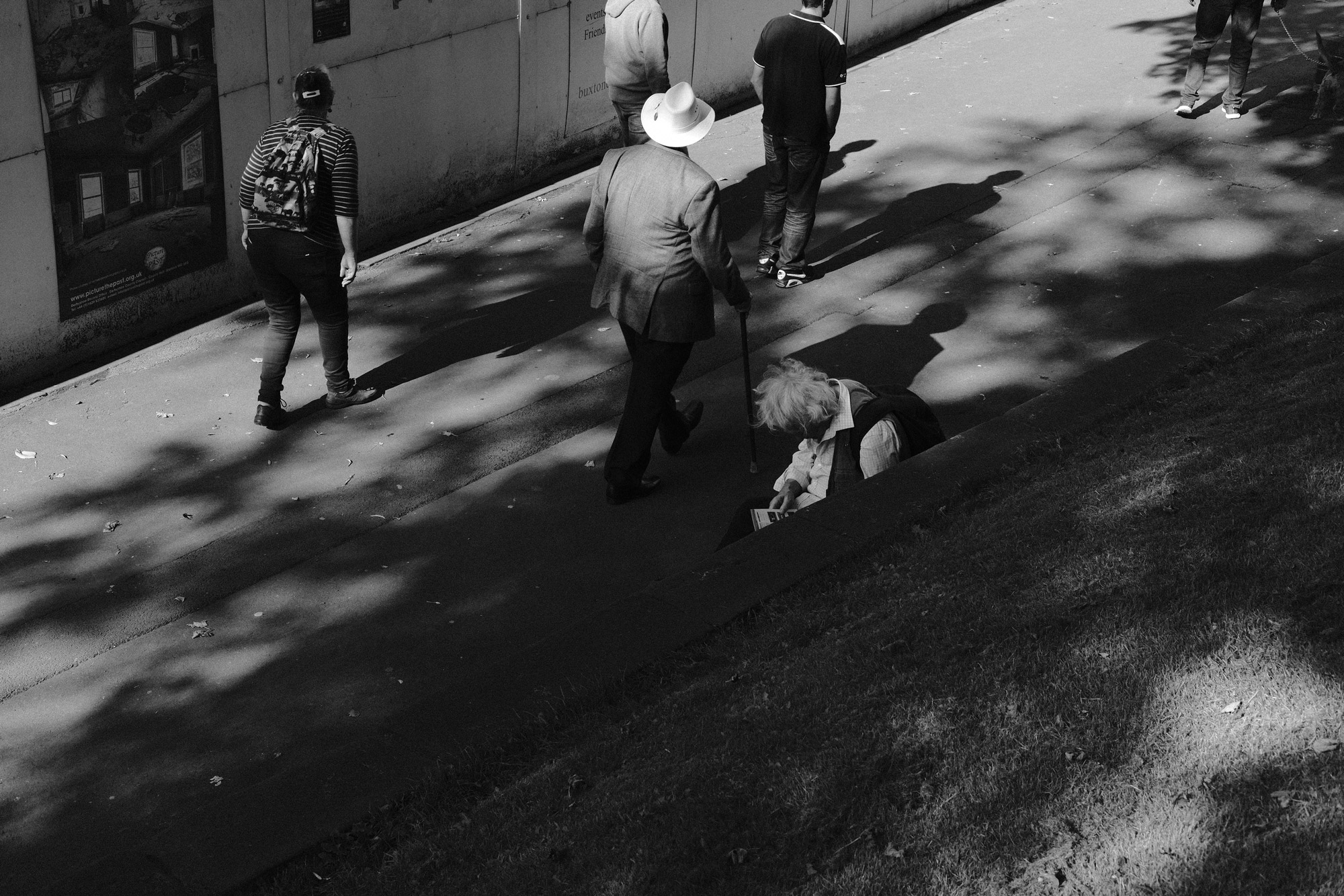 Schwarz-Weiß-Aufnahme, die aus einem hohen Winkel aufgenommen wurde, in der drei Personen auf einer Straße gehen. Ein Mann sitzt fast versteckt. Aufgenommen von Ian Howorth mit der Fujifilm X-Pro3.