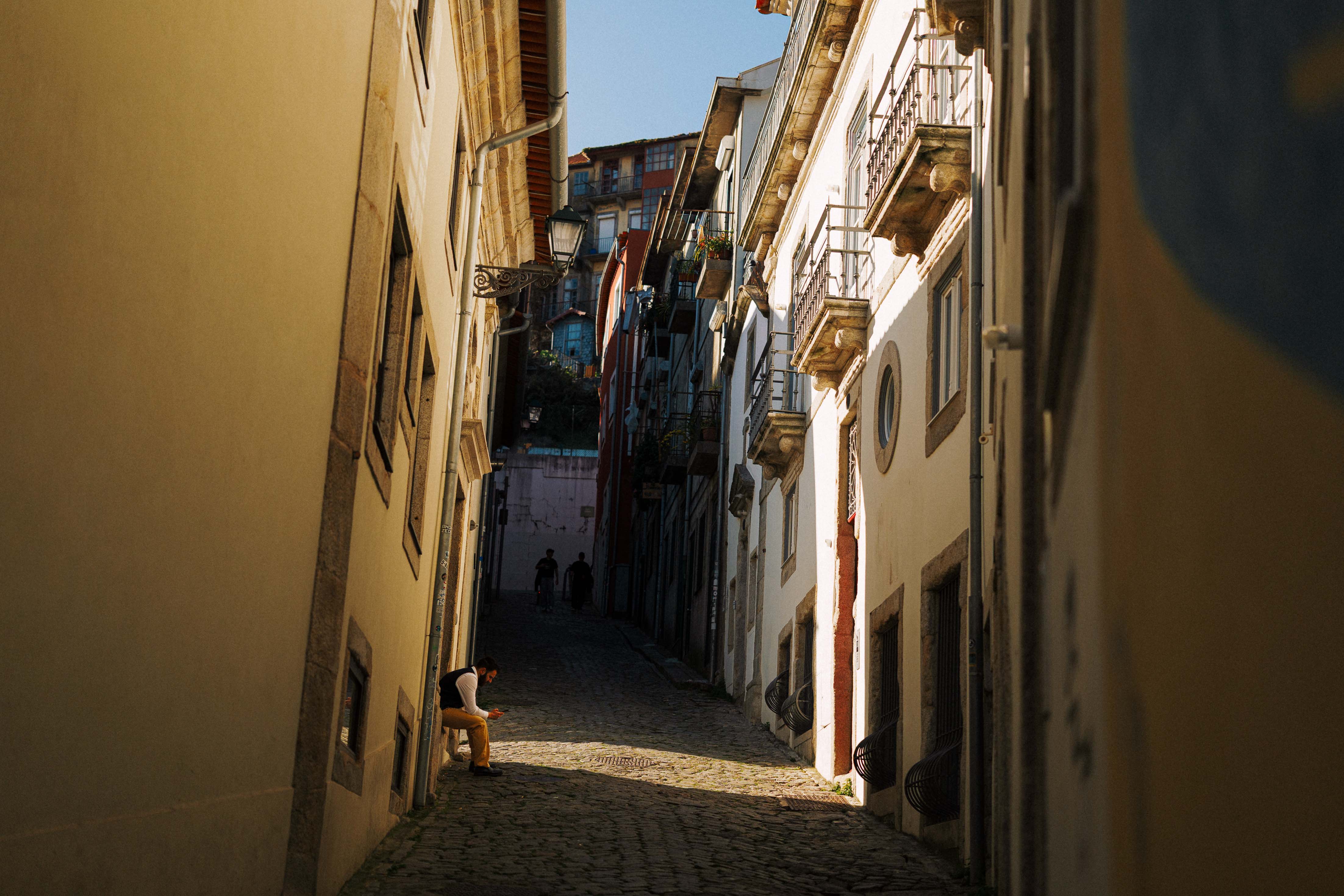 A narrow cobbled street in Porto lined with tall historic buildings, with a single person sitting in a patch of sunlight while the rest of the street falls into shadow.
