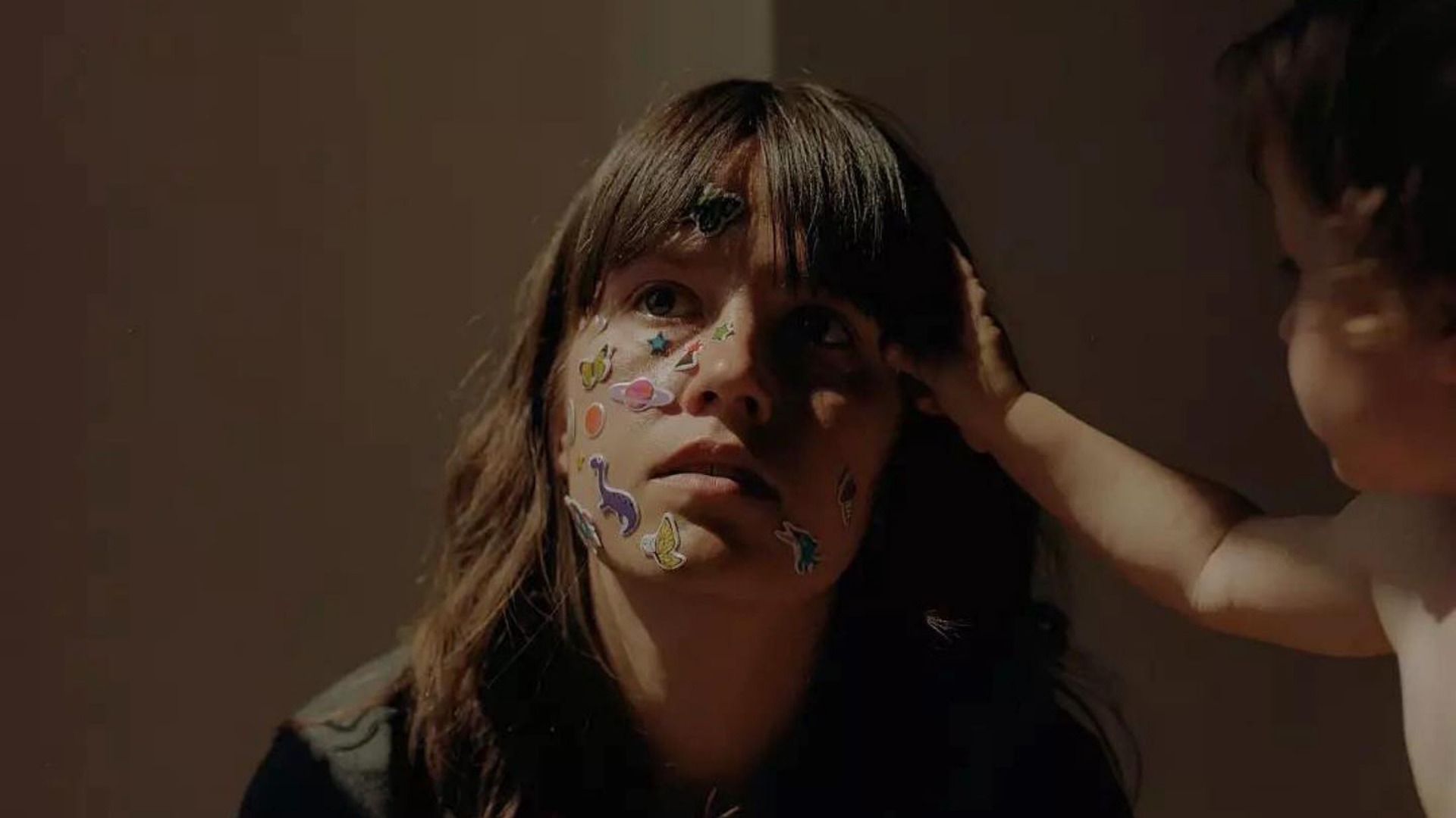 A parent with long brown hair with colourful stickers over their face being touched by a toddler, shot by Tori Ferenc, cropped

