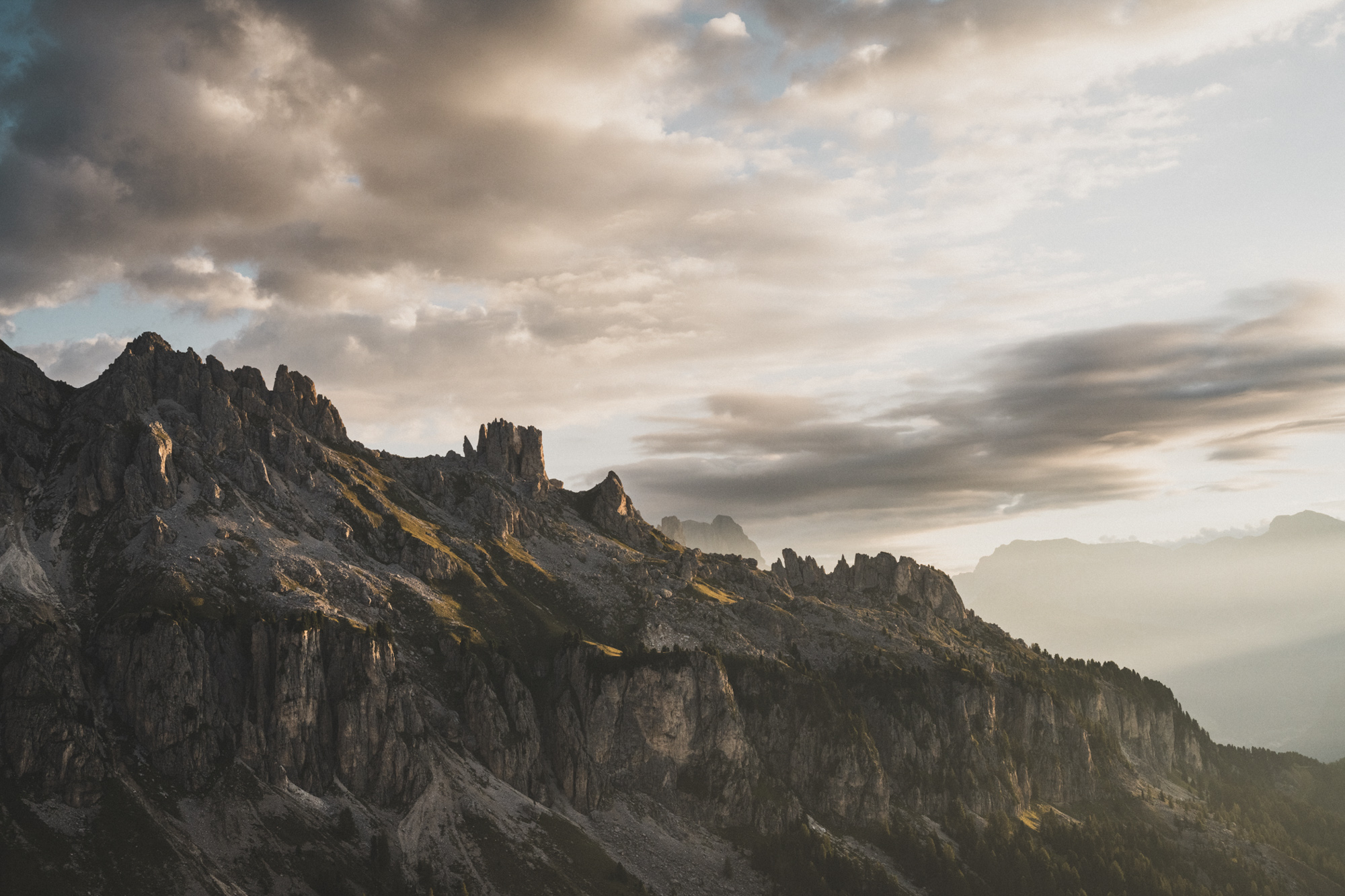 Felsiger Dolomitenkamm bei Sonnenuntergang mit bewölktem Himmel und goldenem Licht, das die senkrechten Felswände beleuchtet.