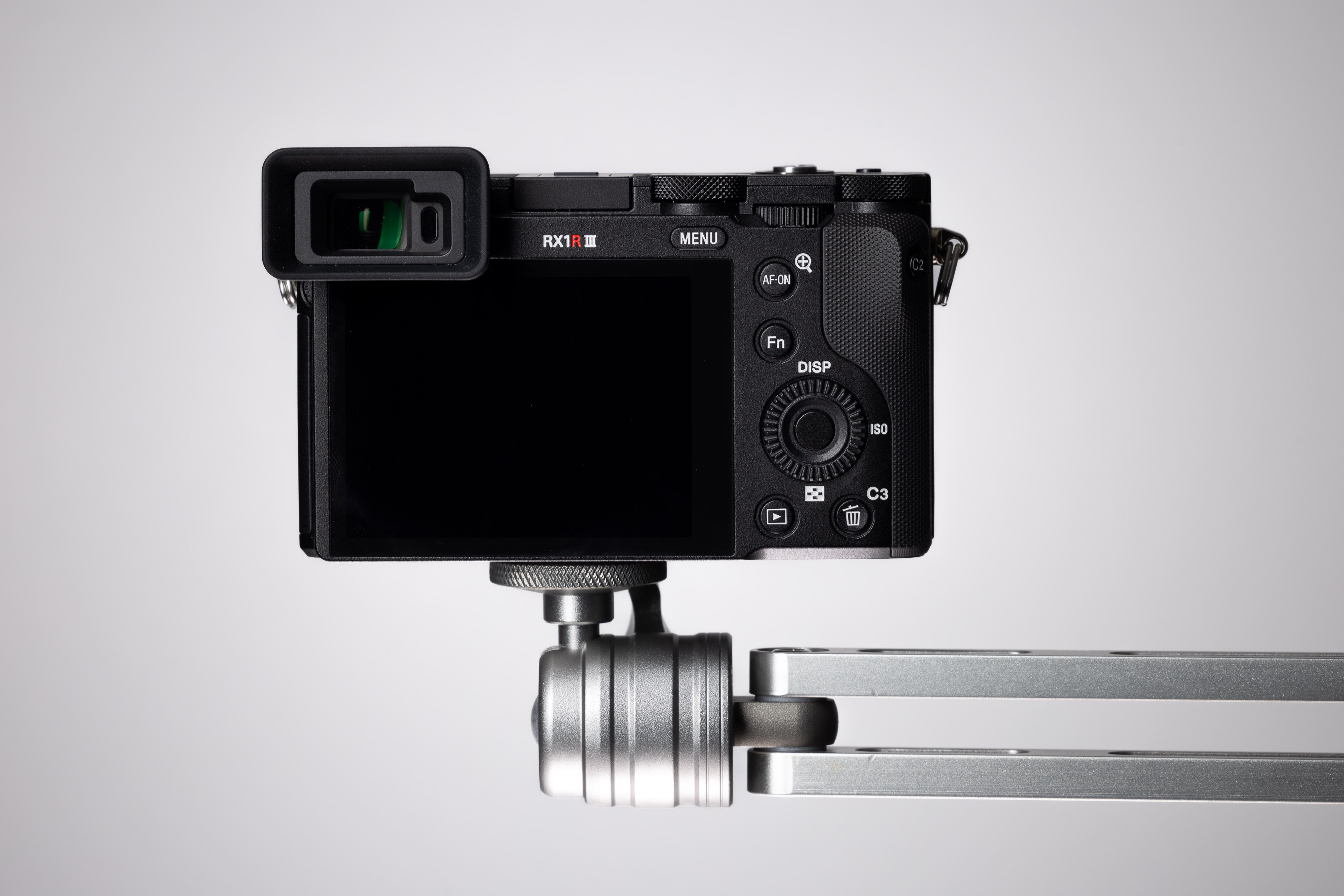 Rückansicht der Sony RX1R III an einem Metallarm, die das fest verbaute hintere LCD, das Steuerrad, die Custom-Tasten und den integrierten elektronischen Sucher vor einem schlichten Studiohintergrund zeigt.