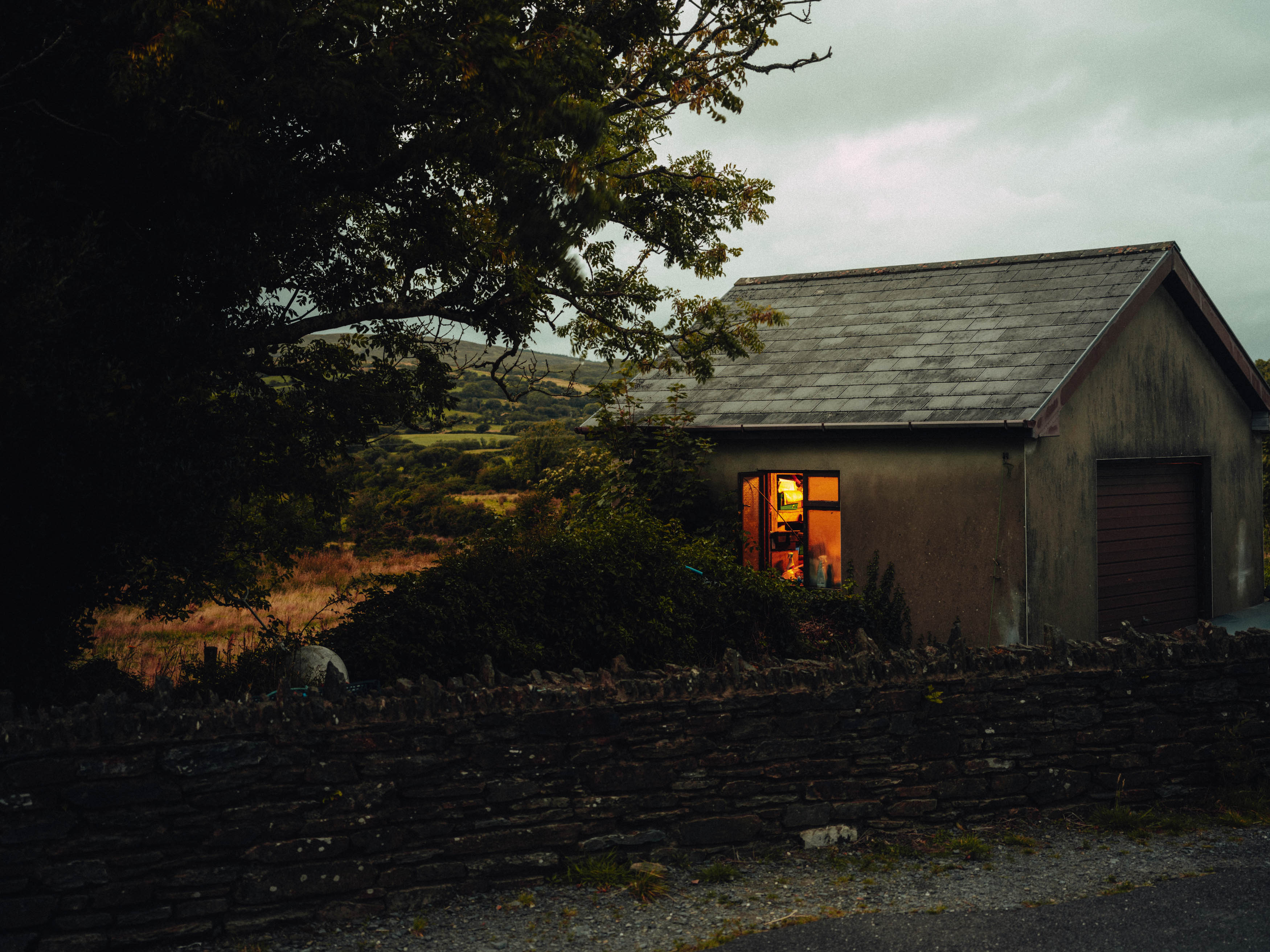 A house and tree behind a stone wall, yellow light filtering through the window, shot by Ian Howorth with a Hasselblad X2D 100C.