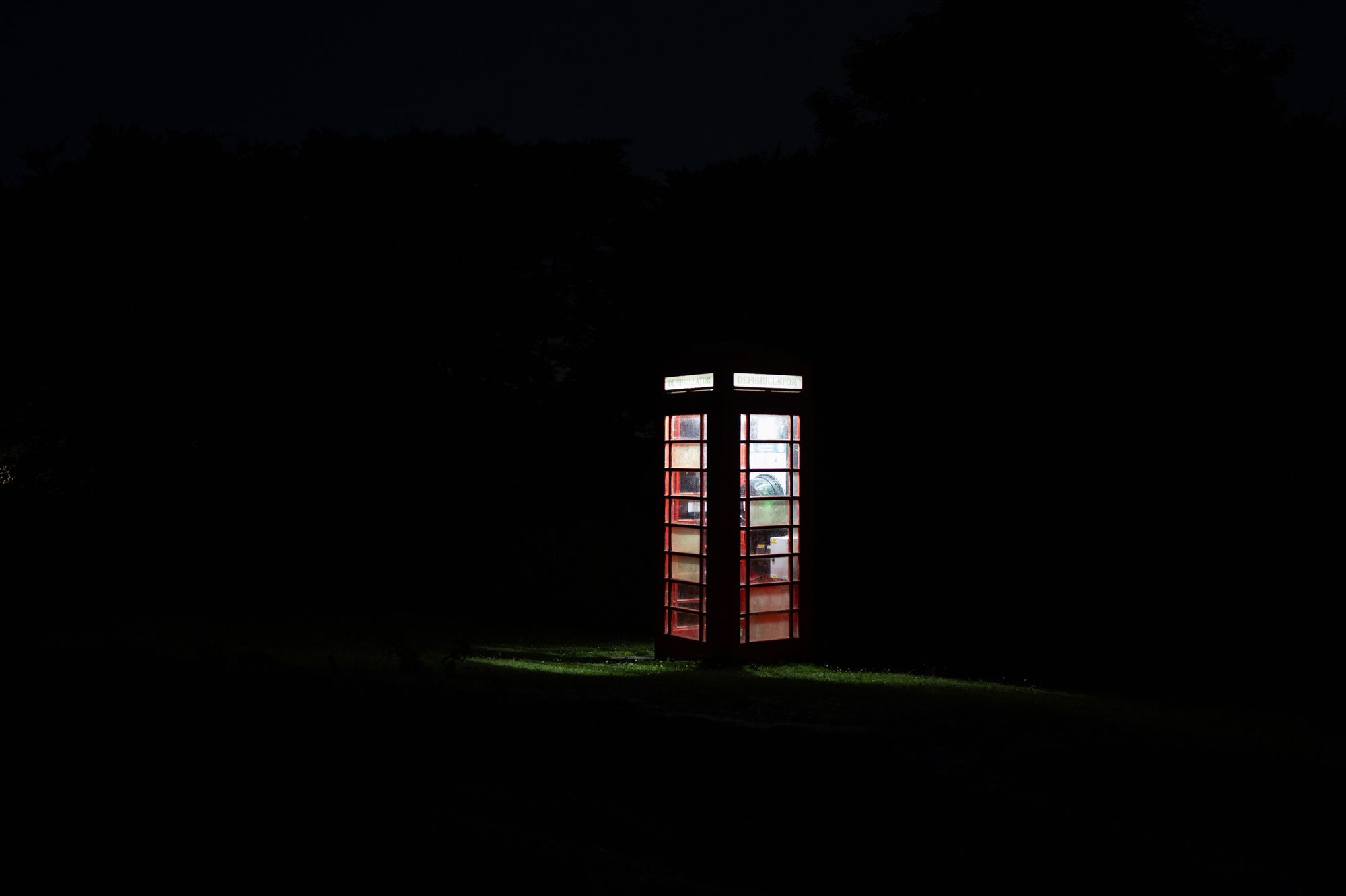 A telephone box with its lights on is shot at night. The light is reflected slightly on the grass around it. Photo by Amy Moore with the Panasonic S1R II.