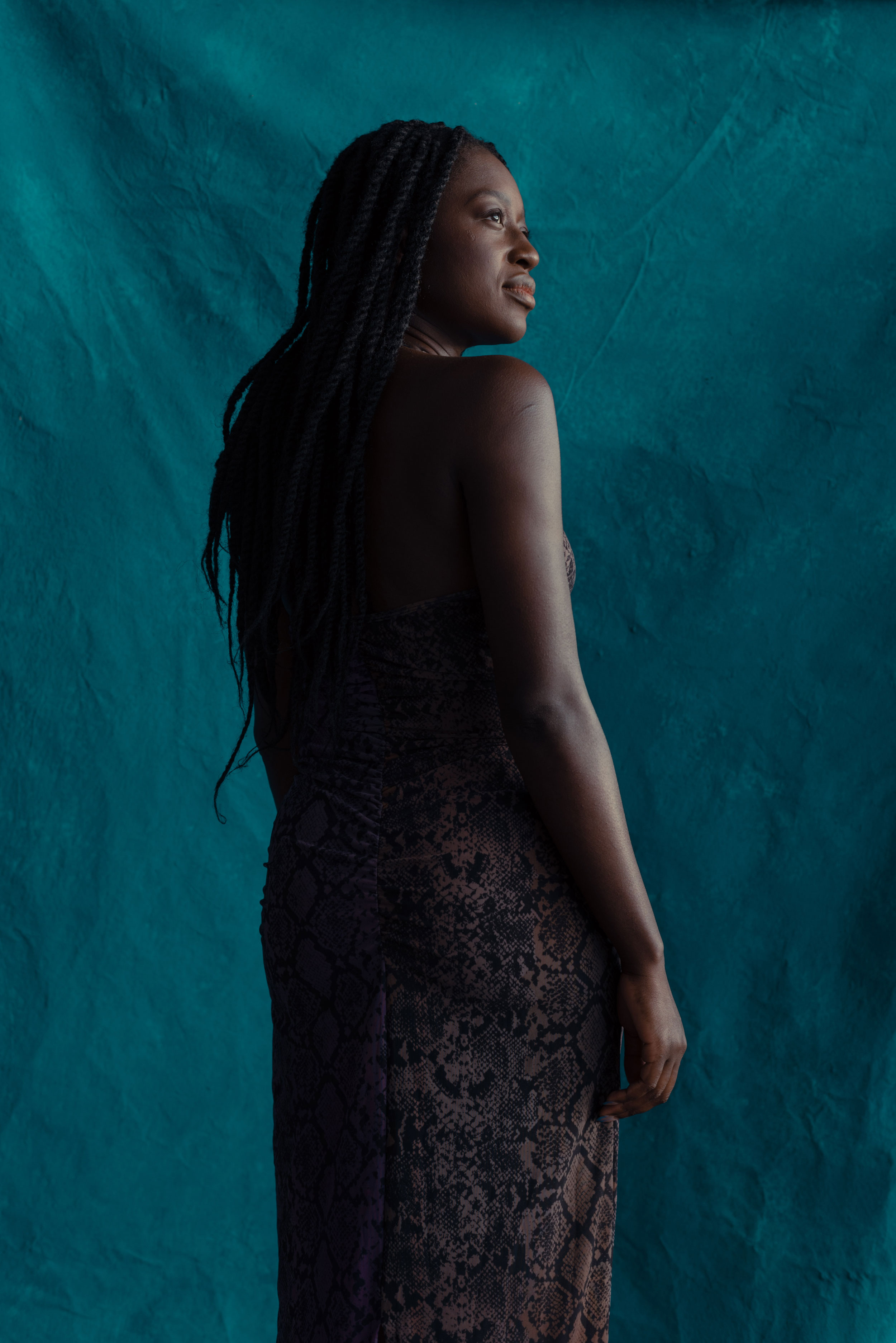 Black woman standing in front of a turquoise wall and looking to the right, photographed from behind, shot by Nicole Benewaah