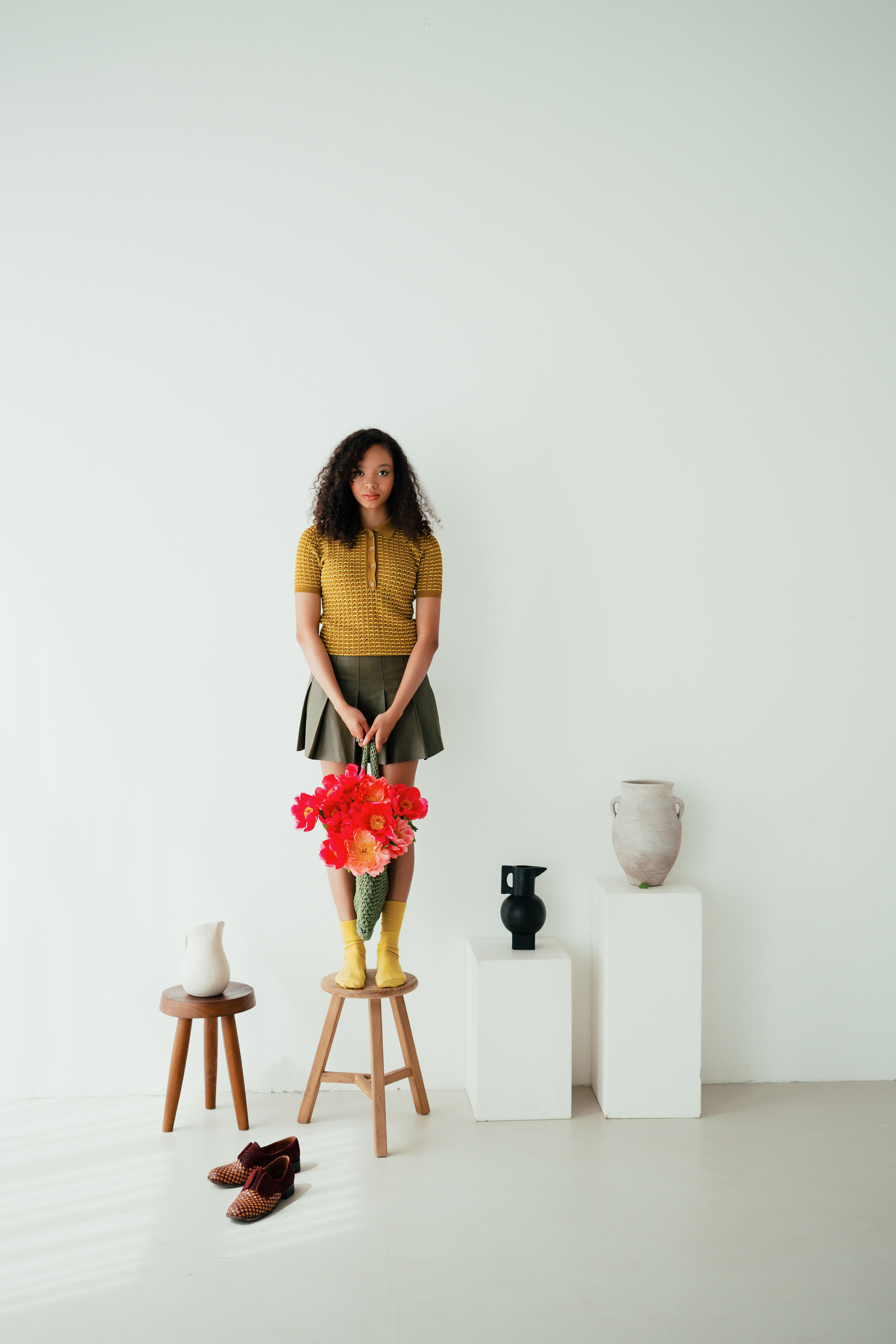 A studio portrait of a model holding vibrant red flowers, standing on a stool surrounded by minimalist ceramics and soft natural light. Shot by Alina Rudya on a Nikon Z6 III.
