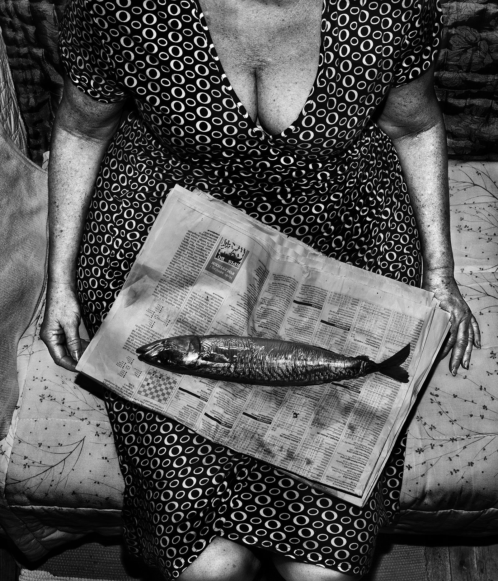Photographie en noir et blanc d'une personne en robe, dont la tête n'est pas visible sur l'image, assise, avec un journal sur les genoux, sur lequel repose un poisson.