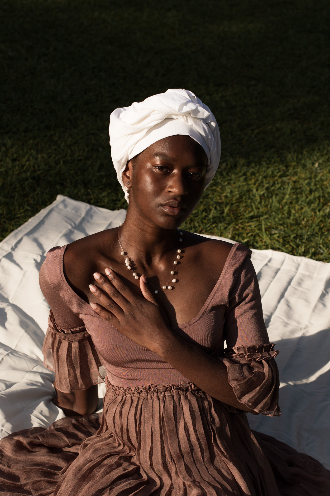 Photo of black woman sitting outside on a picnic blanket wearing a brown dress and white head covering
