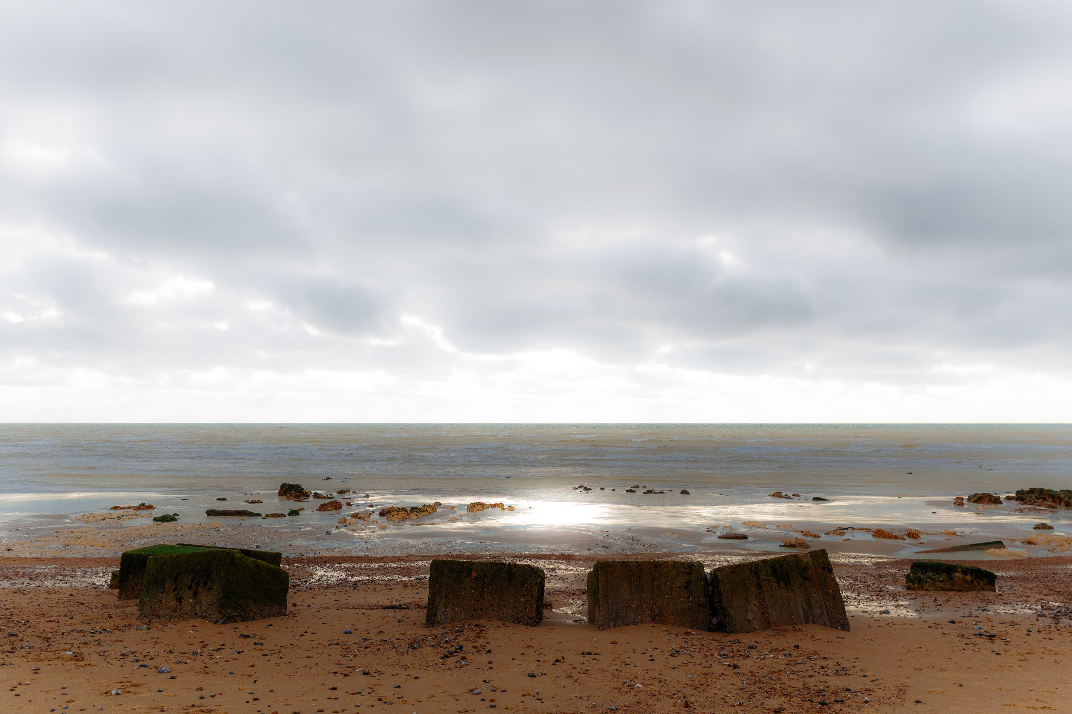 Ein entspanntes Küstenmotiv mit verwitterten Betonwellenbrechern an einem sandigen Ufer bei weichem Tageslicht. Im Hintergrund, das Meer und bewölkter Himmel. Aufgenommen mit einer Ricoh GR IV.