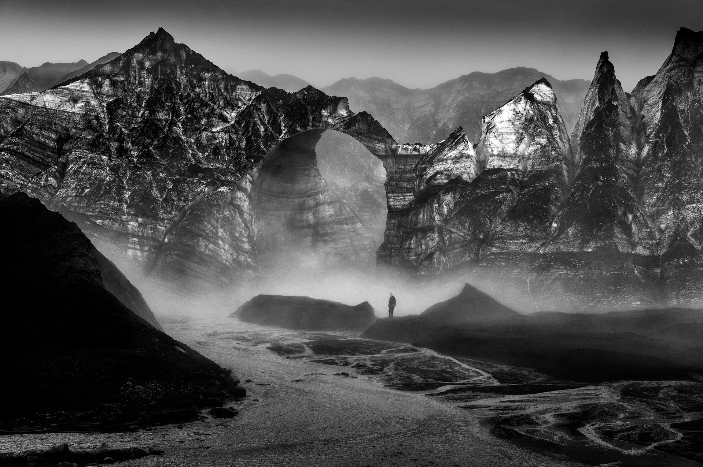 Zwart-wit foto van een berglandschap met een grote boog op de achtergrond, een persoon staat in het midden van de foto, foto gemaakt door Ragnar Axelsson