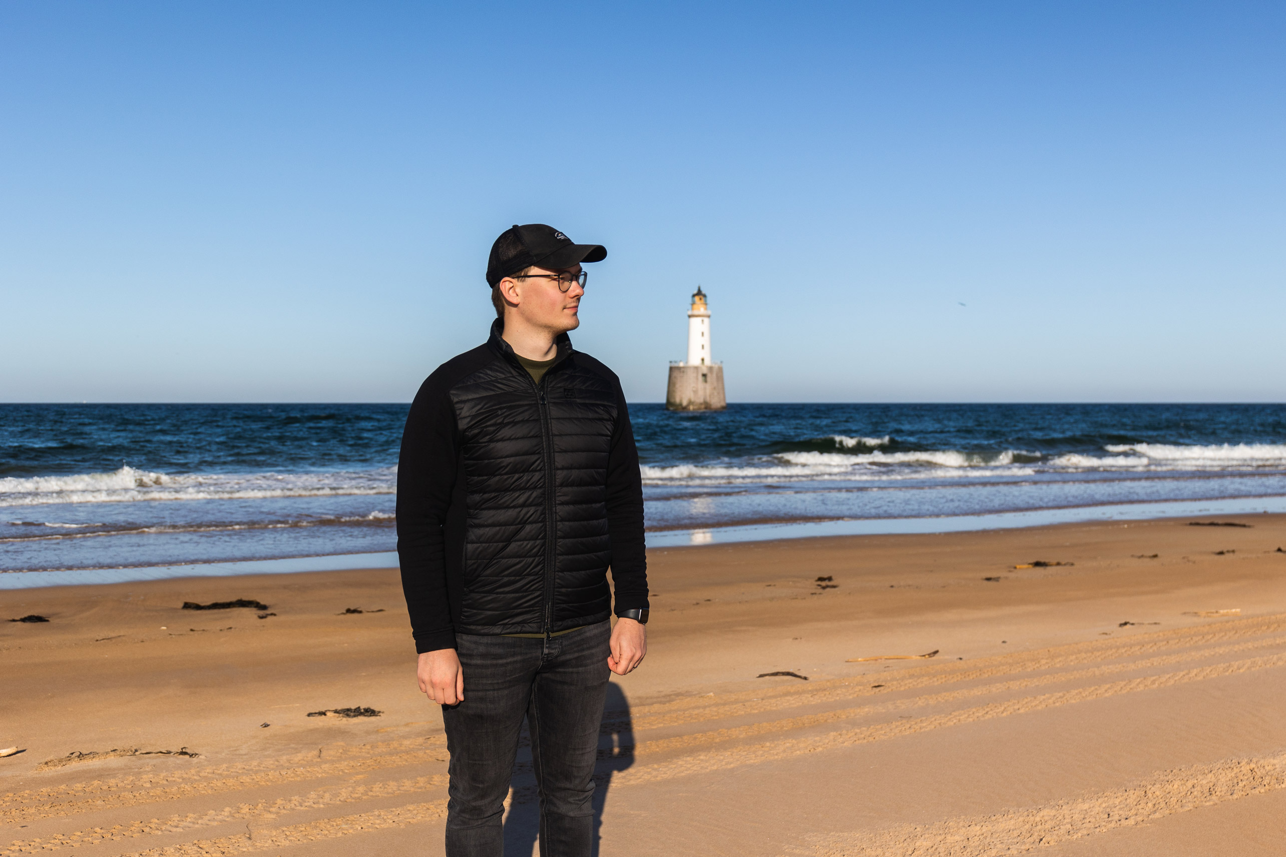 a man wearing a hat standing on a beach with the sea and a lighthouse in the background