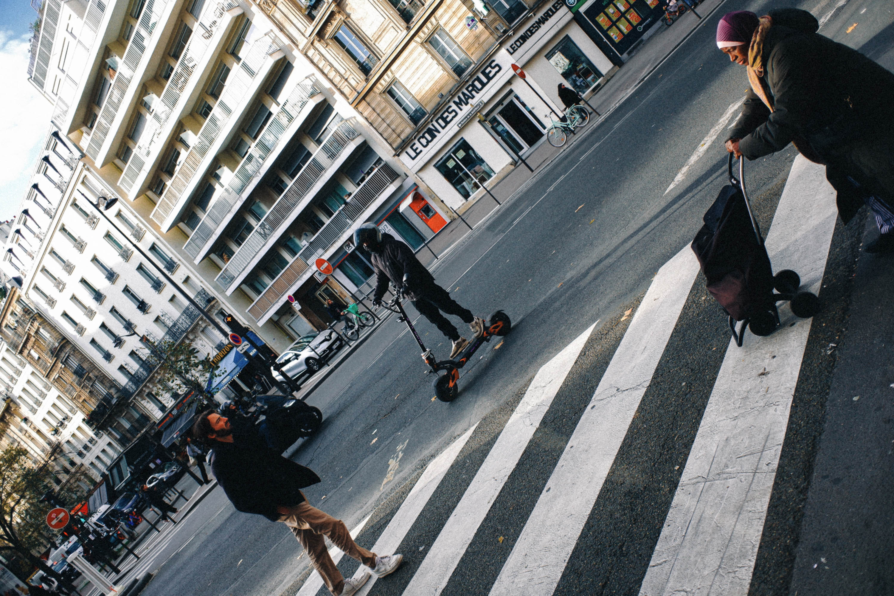 A candid Paris street scene showing several people on a zebra crossing, including a scooter rider, a pedestrian and a person with a shopping trolley. Shot by Wesley Verhoeve on a Fujifilm XF10.