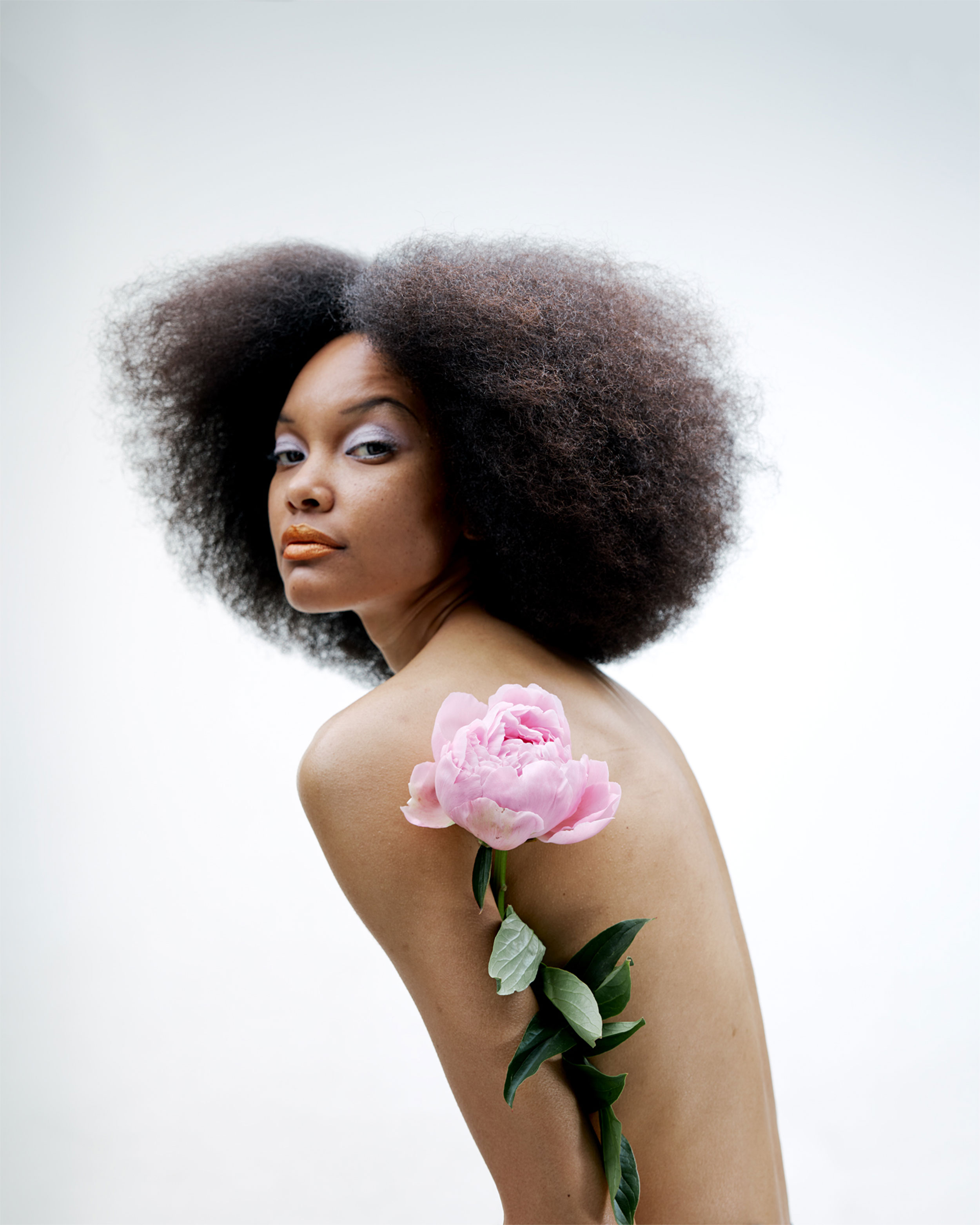 Black woman, topless, photographed from the side with peony between arm and torso, in front of a light background, shot by Lisa Marie Asubonteng