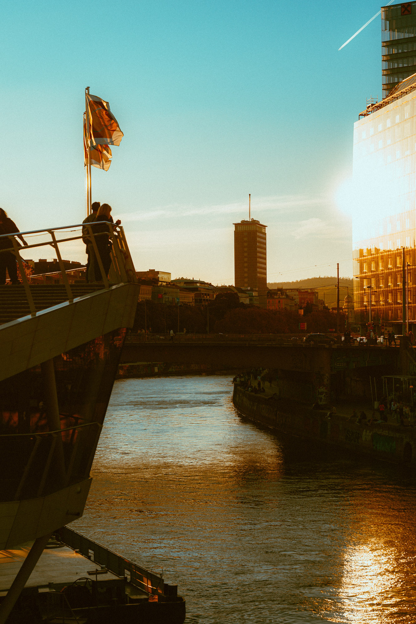 Blick über den Donaukanal bei goldenem Abendlicht: eine Fußgängerbrücke mit Menschen links, reflektierendes Wasser und Gebäude im Hintergrund.