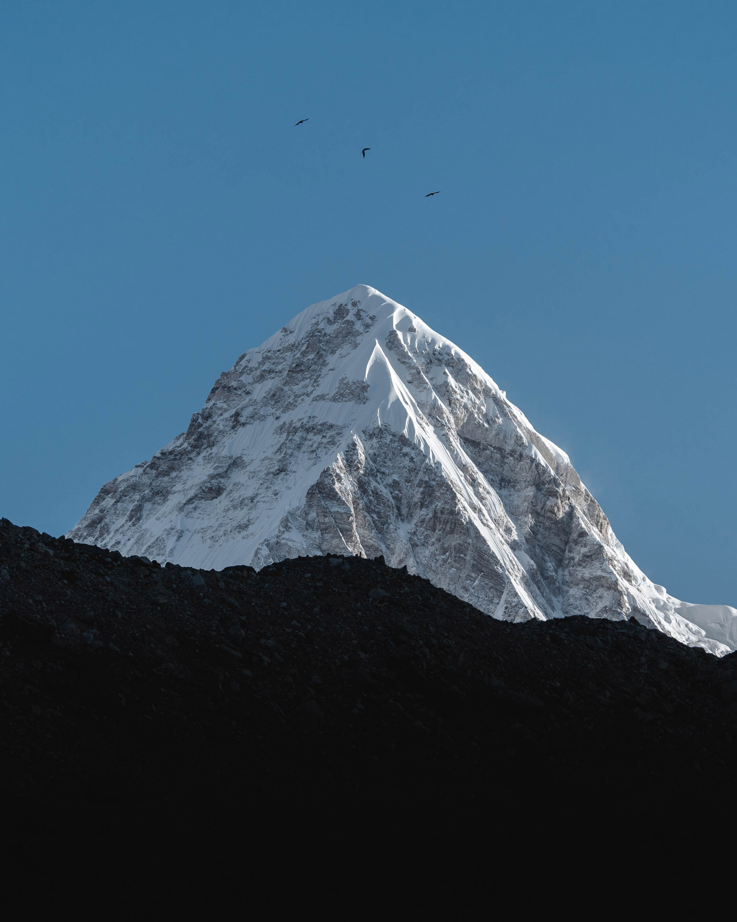 A snowy mountain peak rises above a mountain range in darkness, with blues sky in the background, shot by Jack Anstey with the Sony Alpha A7 IV and the Sony FE 70-200mm f/4 G OSS.