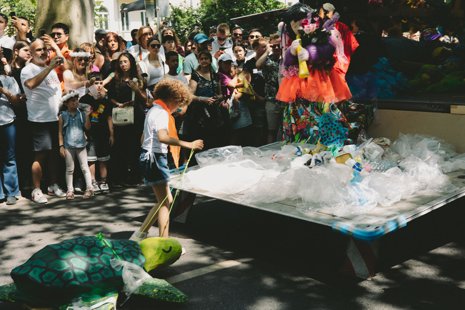 Crowd of people, next to a small platform on which plastic bags lie, child with toy turtle stands in front of it, shot by Stefanie Schmid Rincon with Canon EOS R6