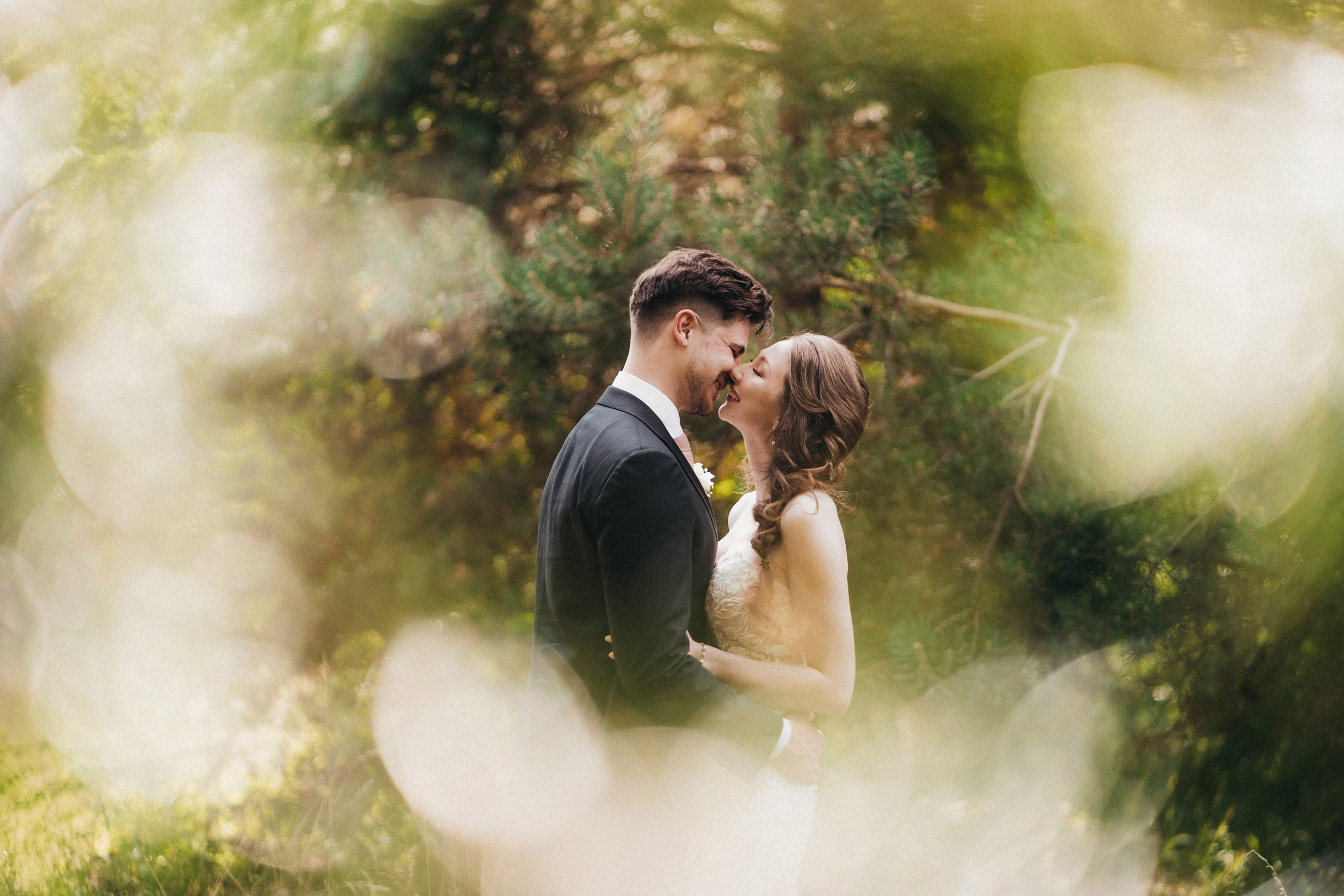 Ein Paar im Anzug und im weißen Hochzeitskleid umarmt sich vor einigen Kiefern, aufgenommen von Hanna Hong mit einer Sony A7 III