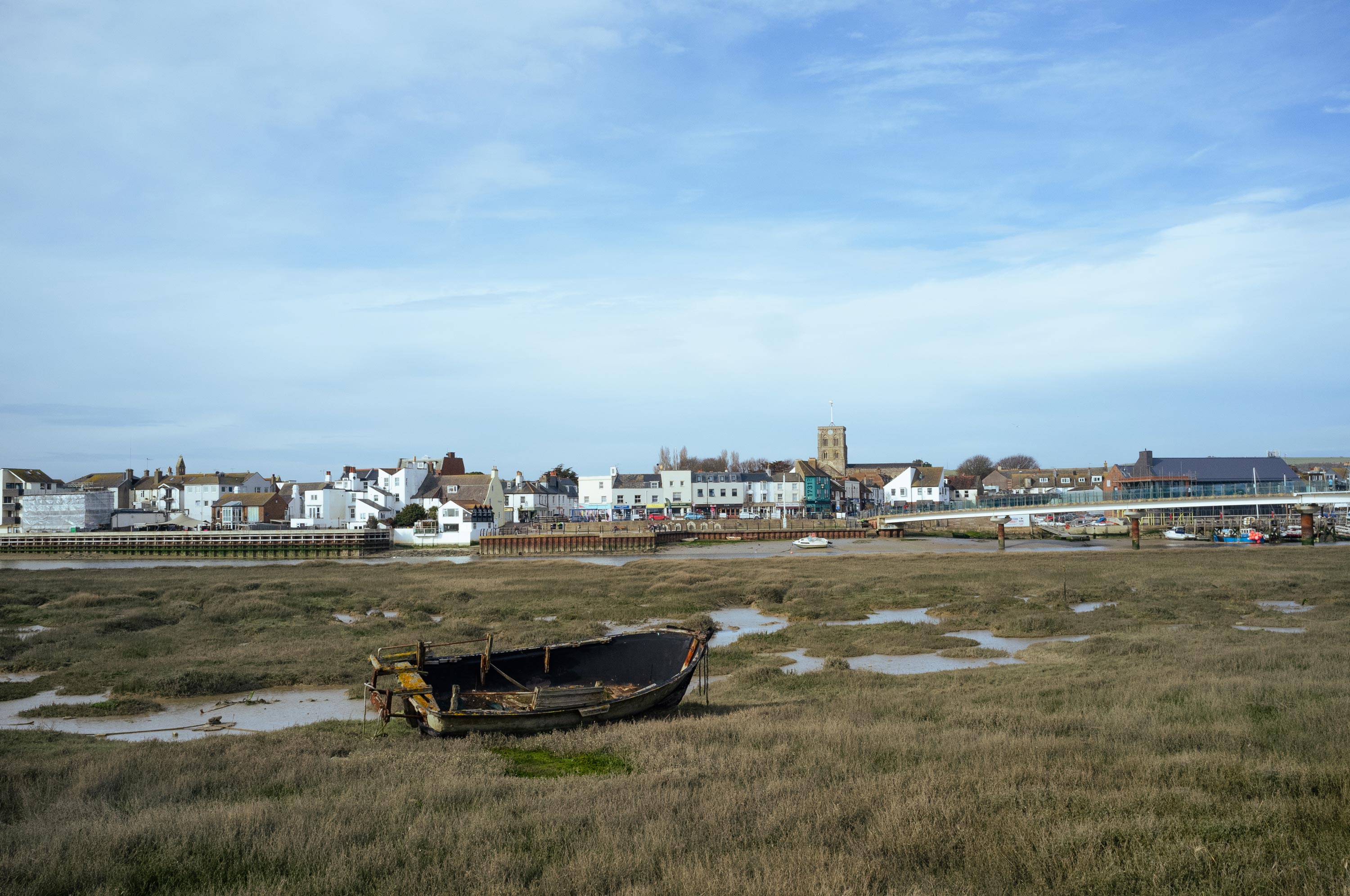 An empty wooden boat sits in the middle of marshy terrain. The tide is low and Shoreham can be seen in the distance.