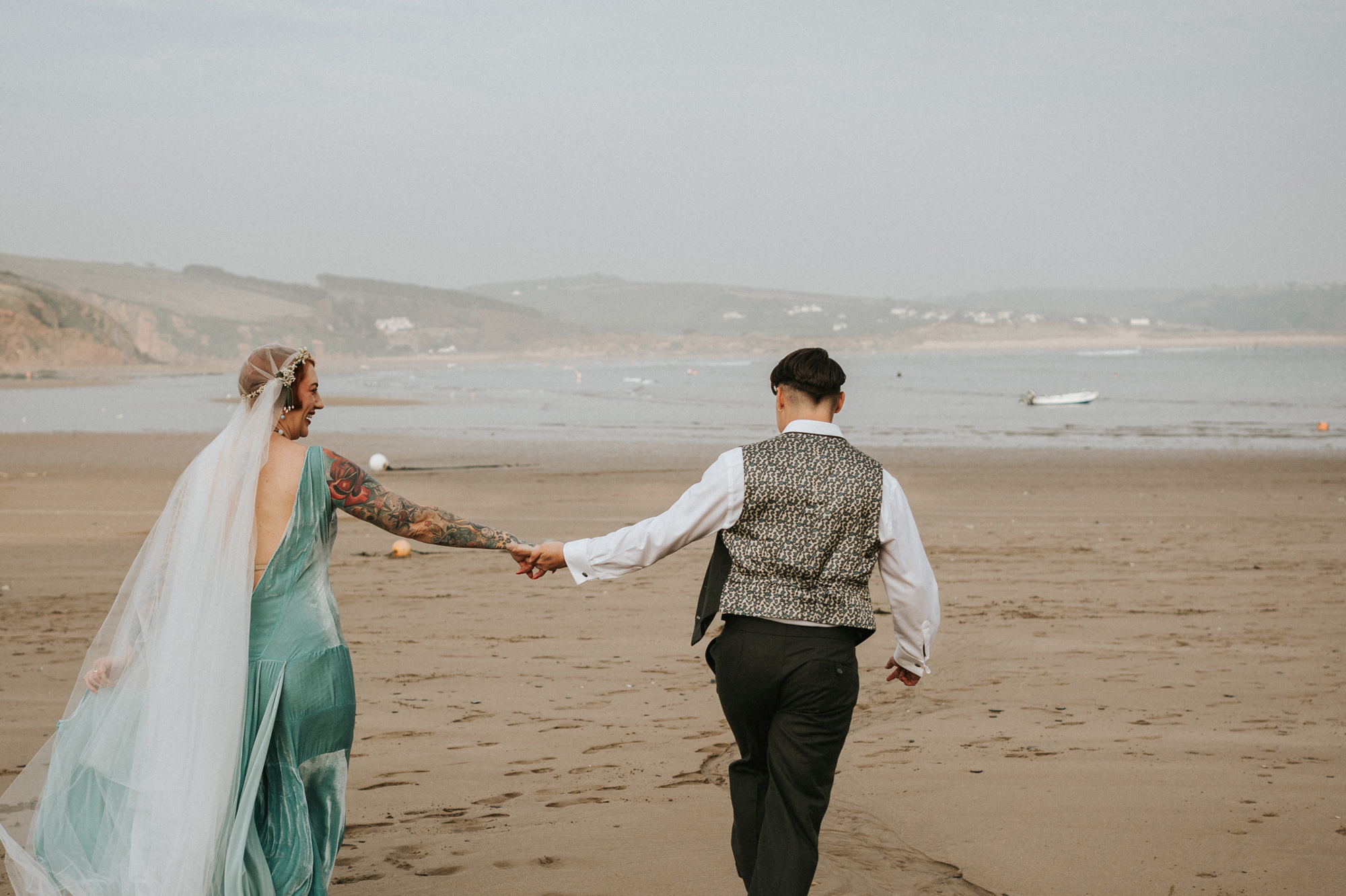 Newlyweds holding hands while walking along the beach. Untraditional wedding gown, green colour, with a long veil. Shot by Steph Large