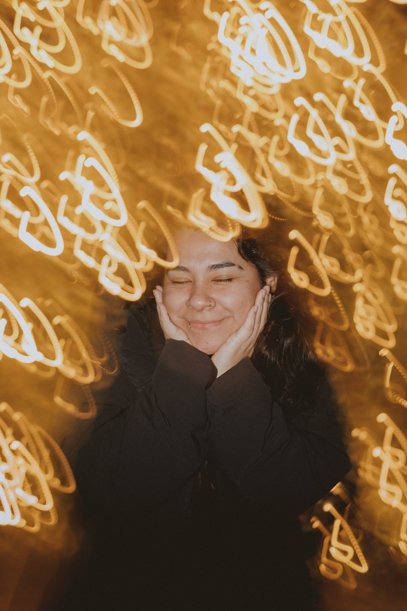 Creative portrait of a smiling woman framed by warm golden light trails and bokeh, creating a dreamy atmosphere with long exposure photography effects.