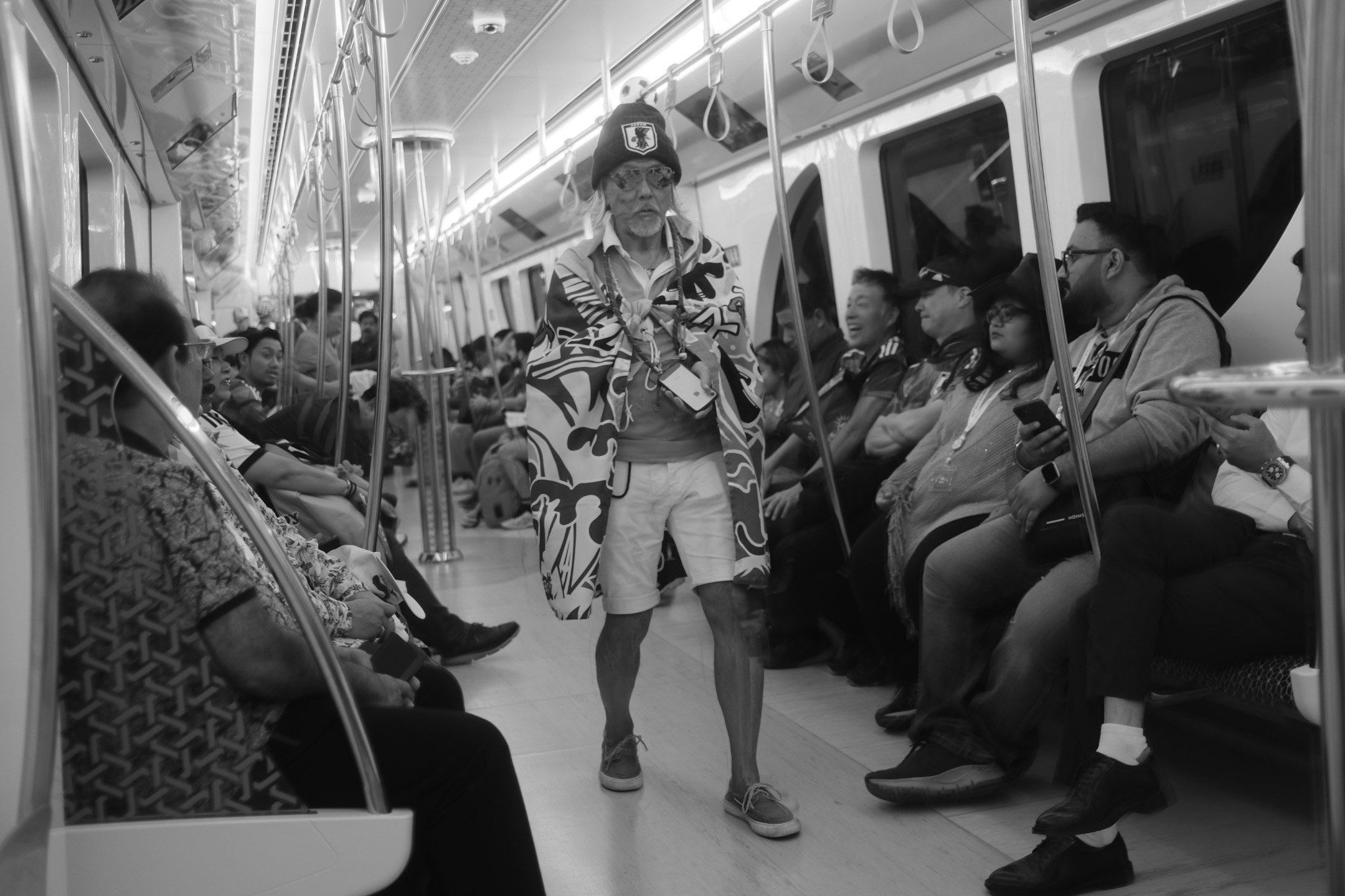 Photo en noir et blanc d’un supporter japonais portant un chapeau, des lunettes de soleil et un short, enveloppé dans un drapeau, marchant dans les transports en commun pendant la Coupe du monde 2022, prise par Ryu Voelkel avec un Fujifilm X100V