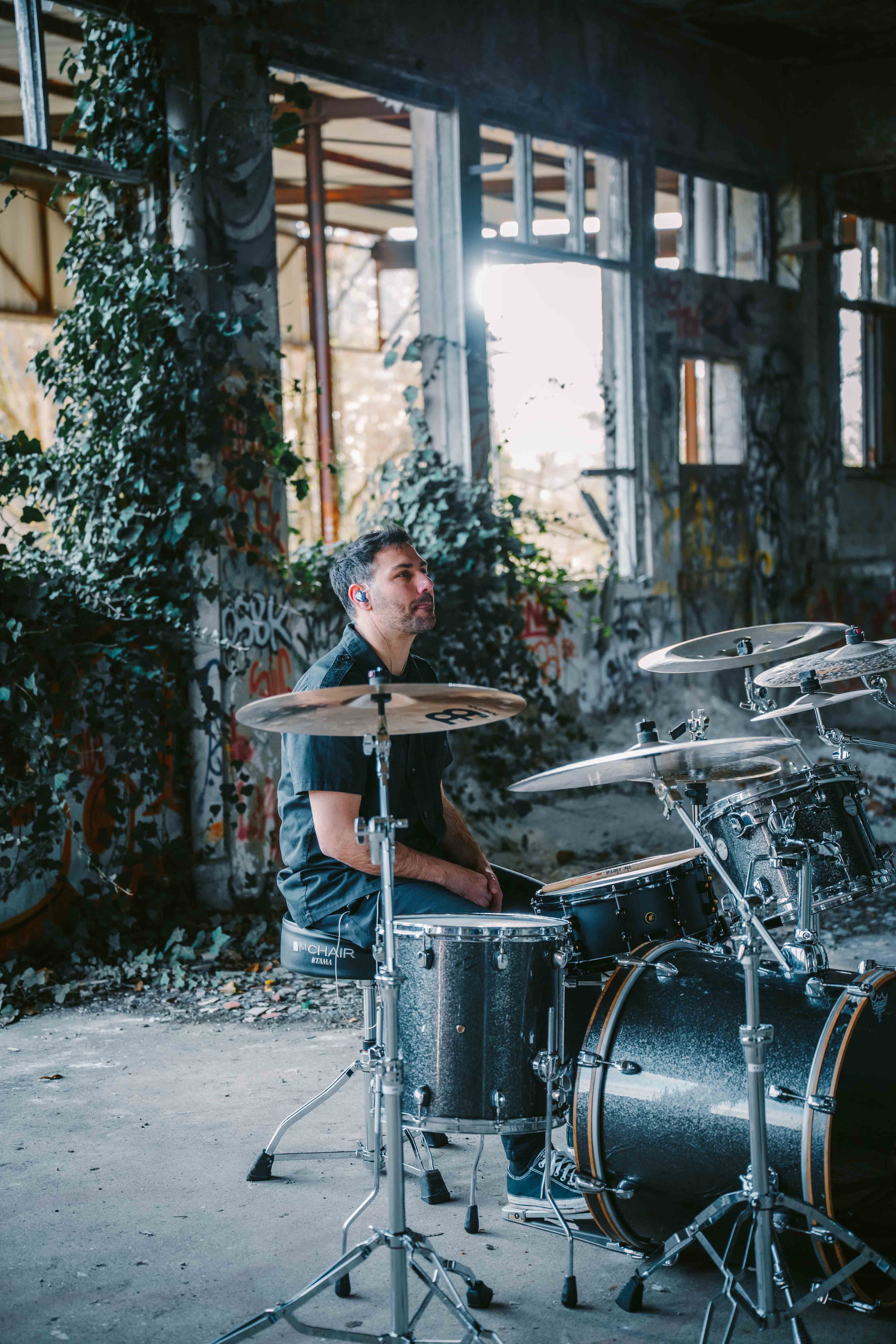A drummer sits behind a black drum kit inside an abandoned, graffiti-covered building, surrounded by overgrown plants and natural light streaming through broken windows. Photo taken by Lukas Davos.