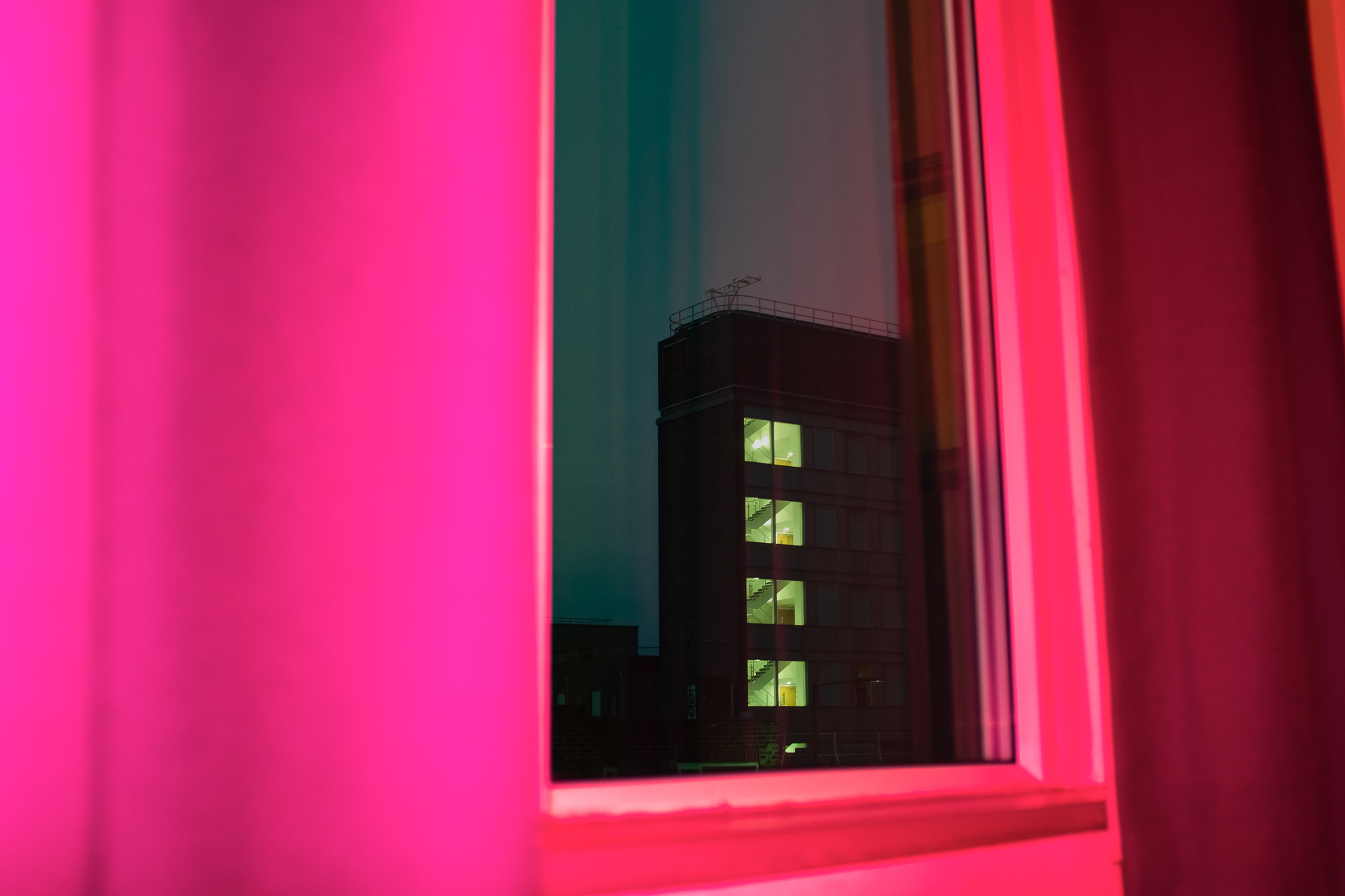 Een flatgebouw bij nacht omlijst door een raam dat baadt in een roze licht, gefotografeerd door Ian Howorth.