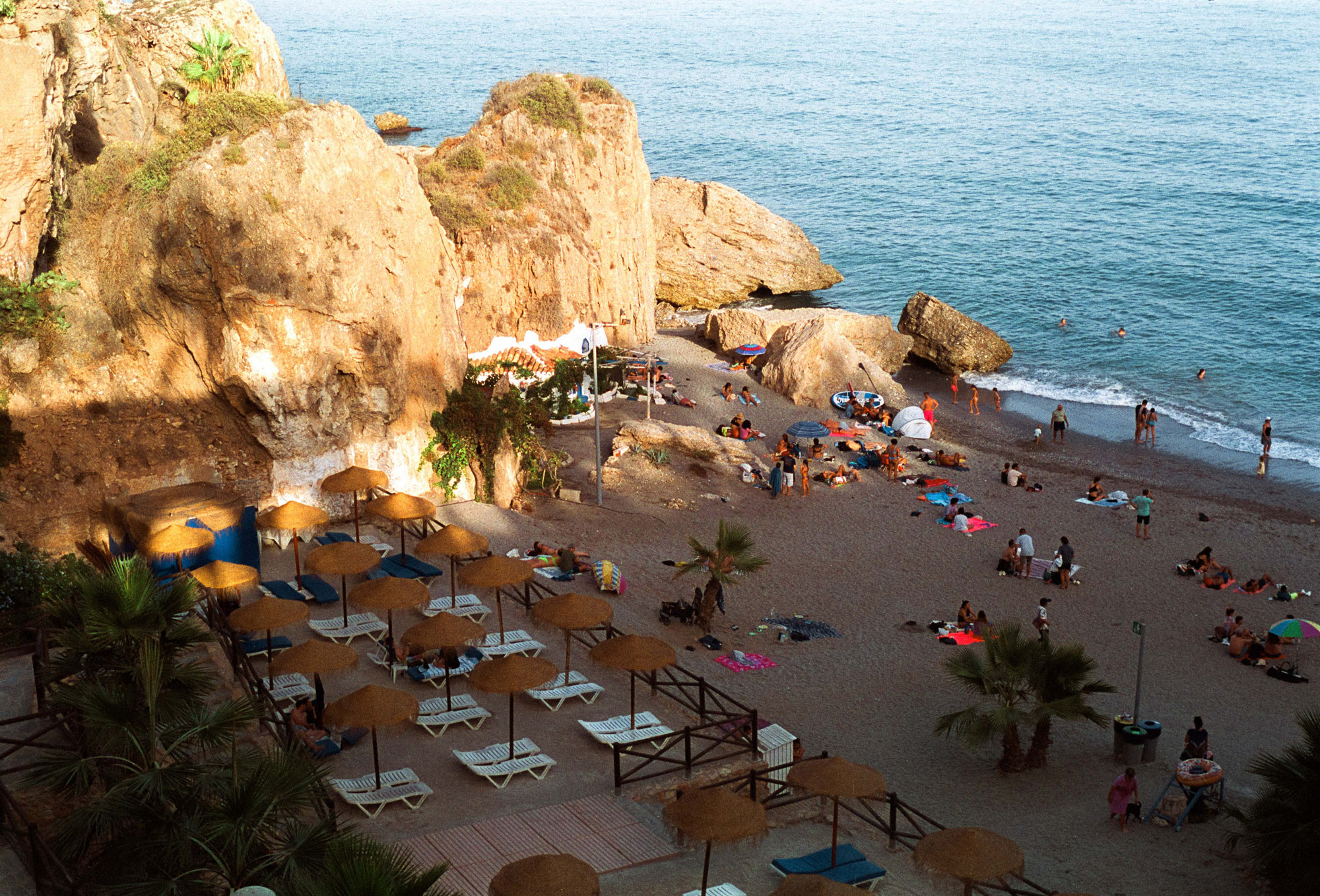 A small beach enclave surrounded by rocky crags with people lying down on sunbeds and enjoying the sea. By Ian Howorth.