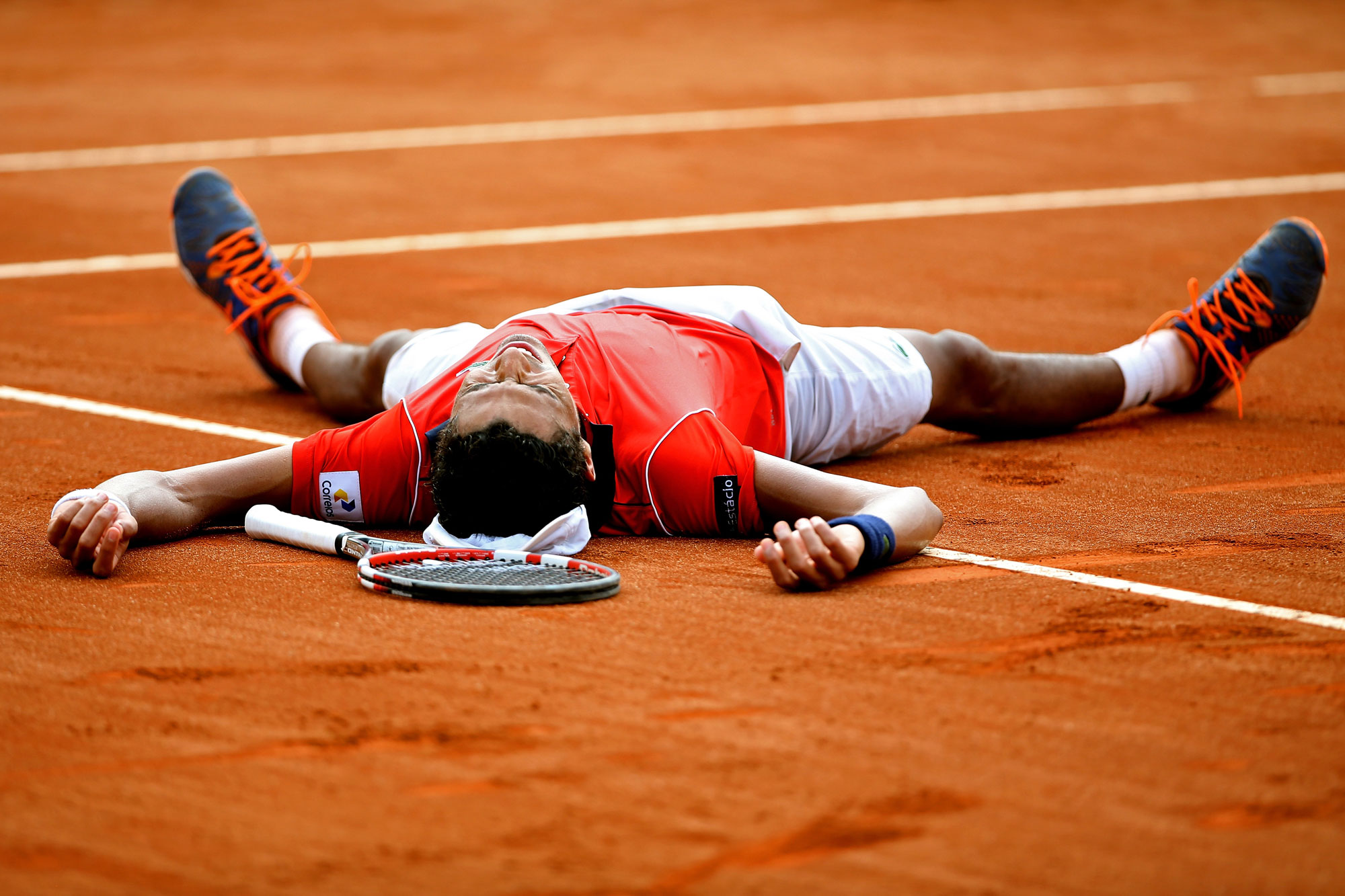 Thiago Monteiro of Brazil celebrates a match point against Jo-Wilfried Tsonga of France during the Rio Open at Jockey Club Brasileiro in Rio de Janeiro, Brazil. He is lying on the clay floor on his back with his arms and legs outstretched, his racquet in front of him. 