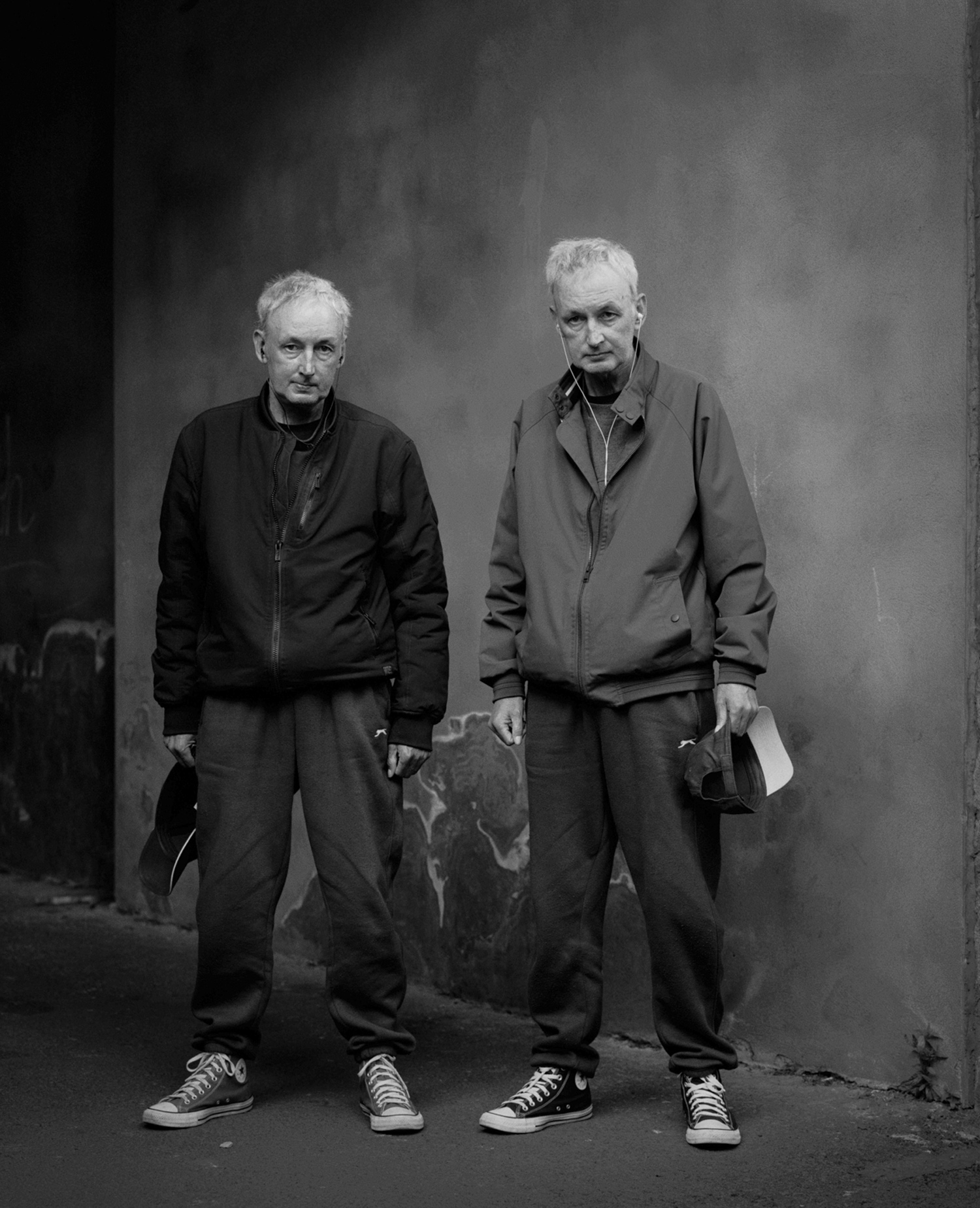 Une photo de rue en noir et blanc d'une paire de jumeaux âgés à l'extérieur, tenant tous deux leurs casquettes dans des mains opposées, photo de Simon Murphy