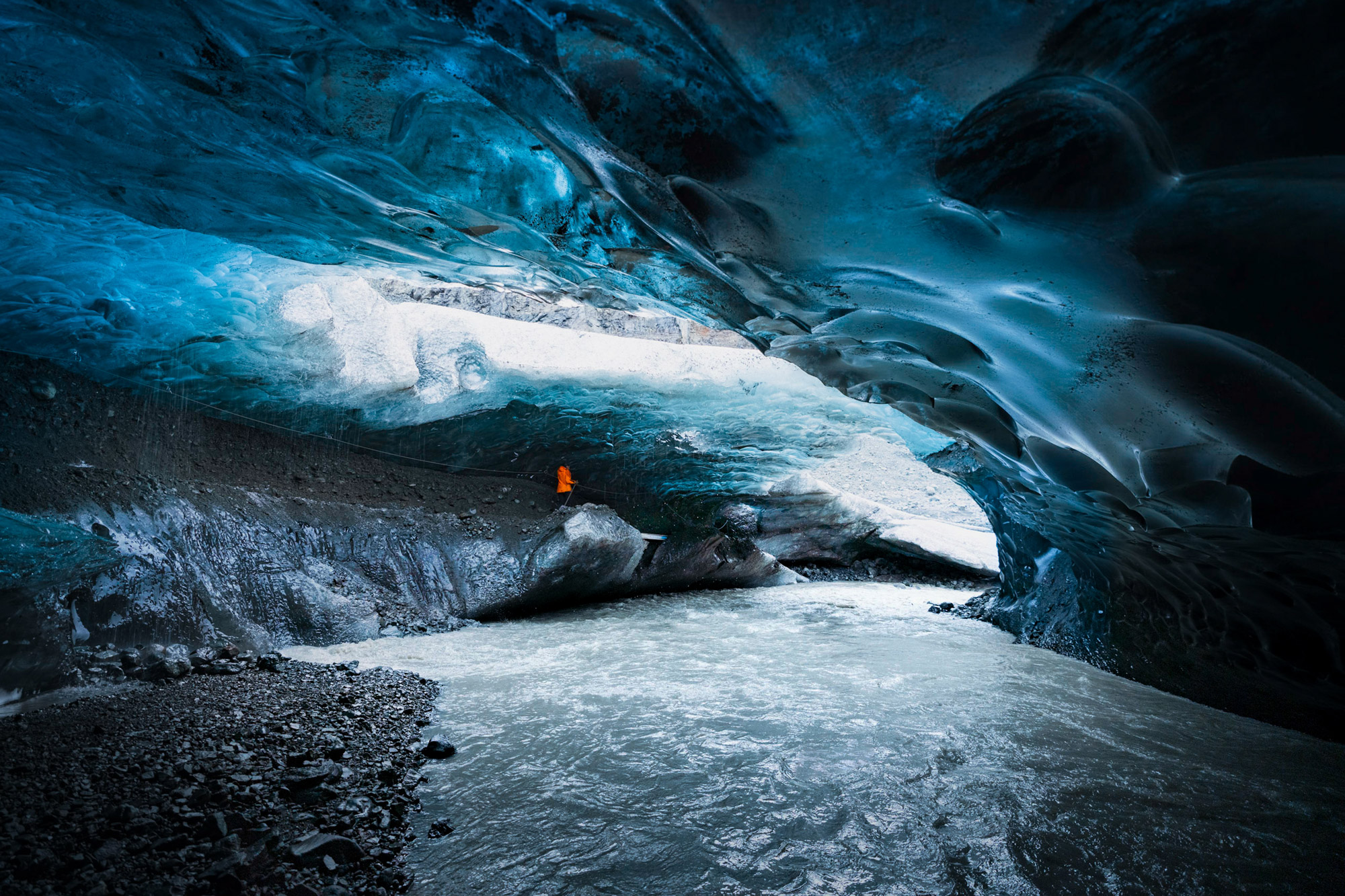 Gewässer in einer riesigen Gletscherhöhle, Person mit oranger Jacke im Hintergrund auf einem Felsen