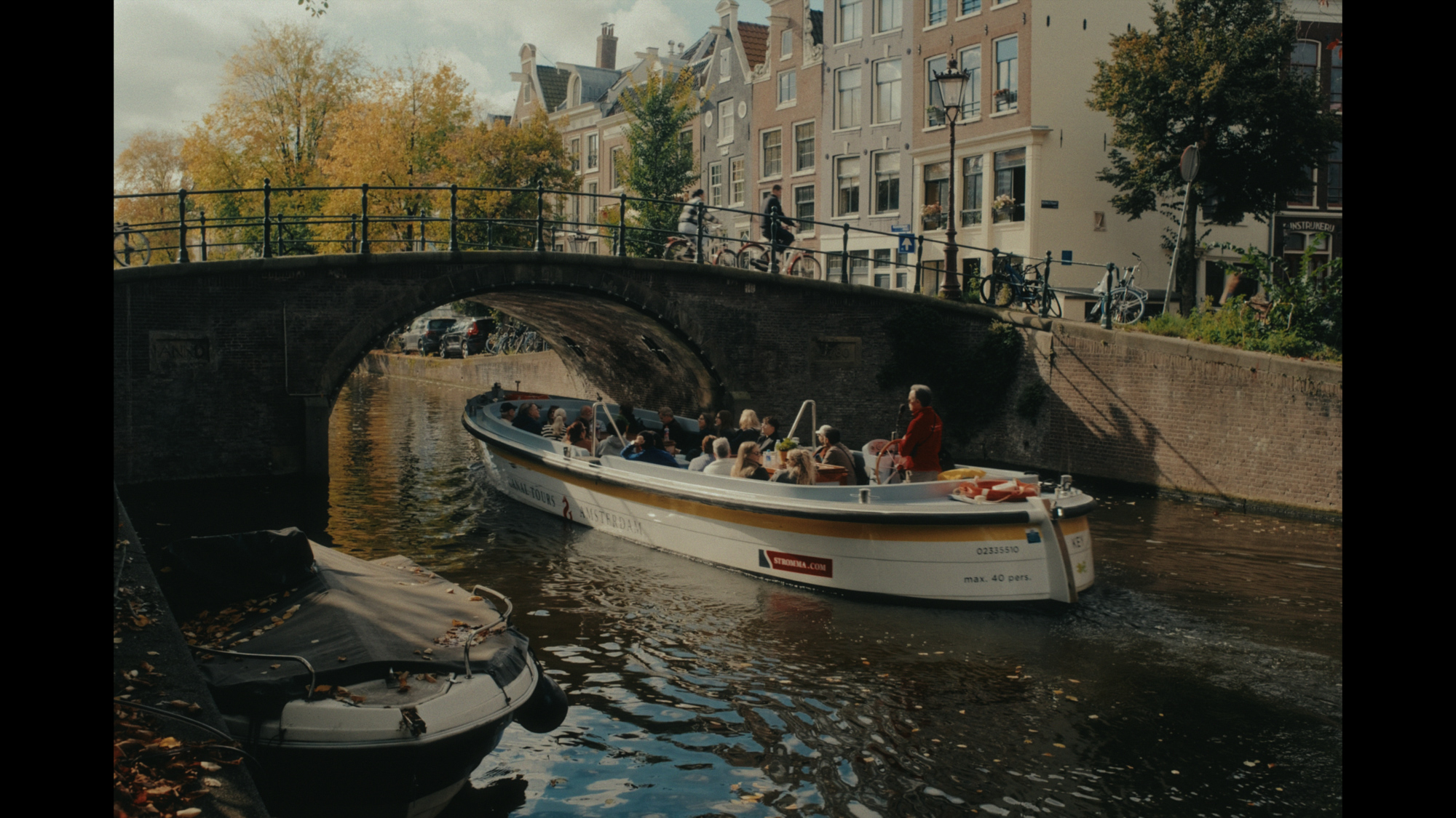 A tour boat filled with passengers navigates a picturesque canal in Amsterdam beneath a classic arched stone bridge.