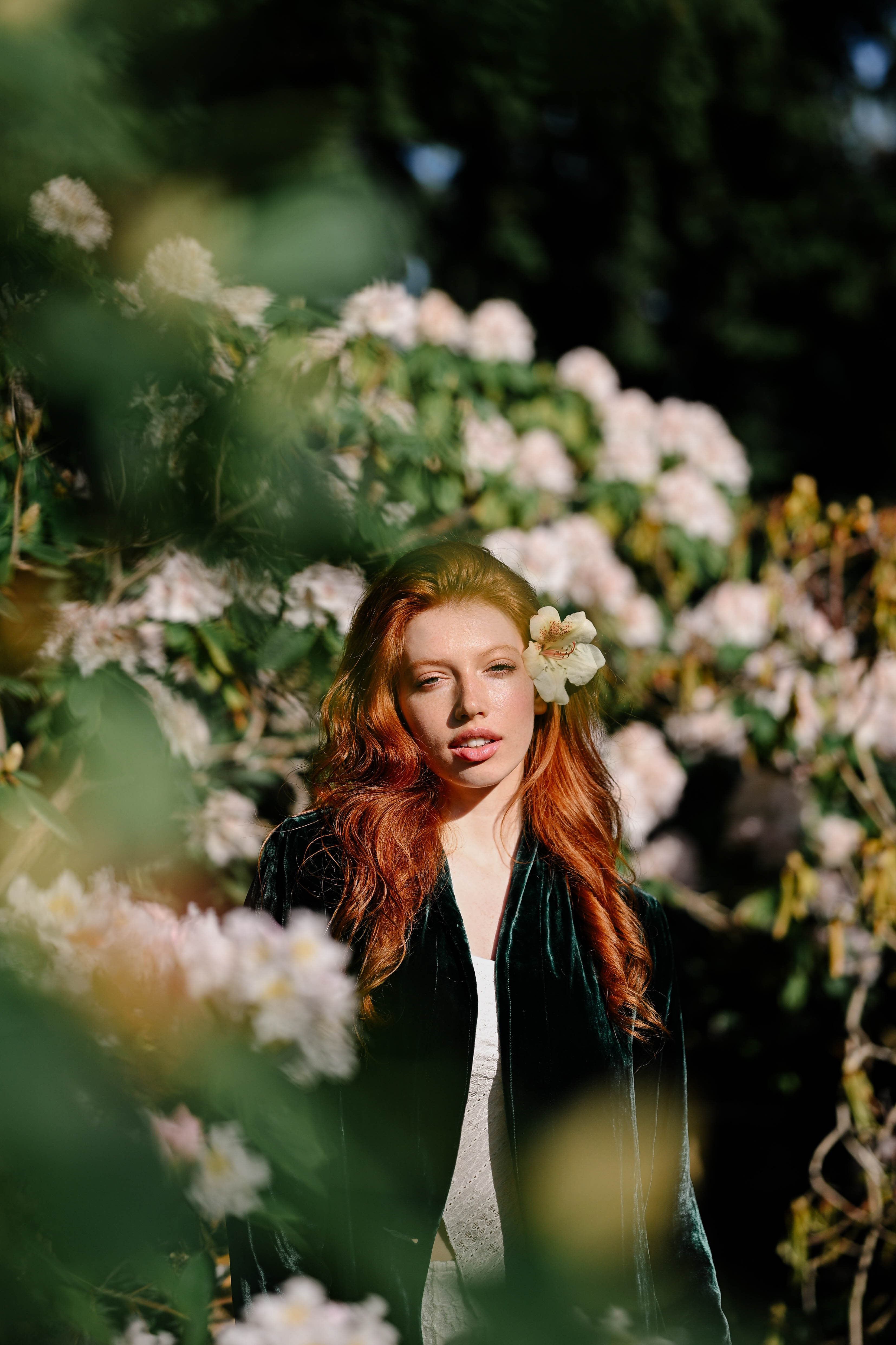 A red-haired model wearing a green velvet jacket and a white flower in their hair, surrounded by blooming bushes and soft morning light in Berlin’s Humboldthain park. Shot by Alina Rudya on a Nikon Z6 III.