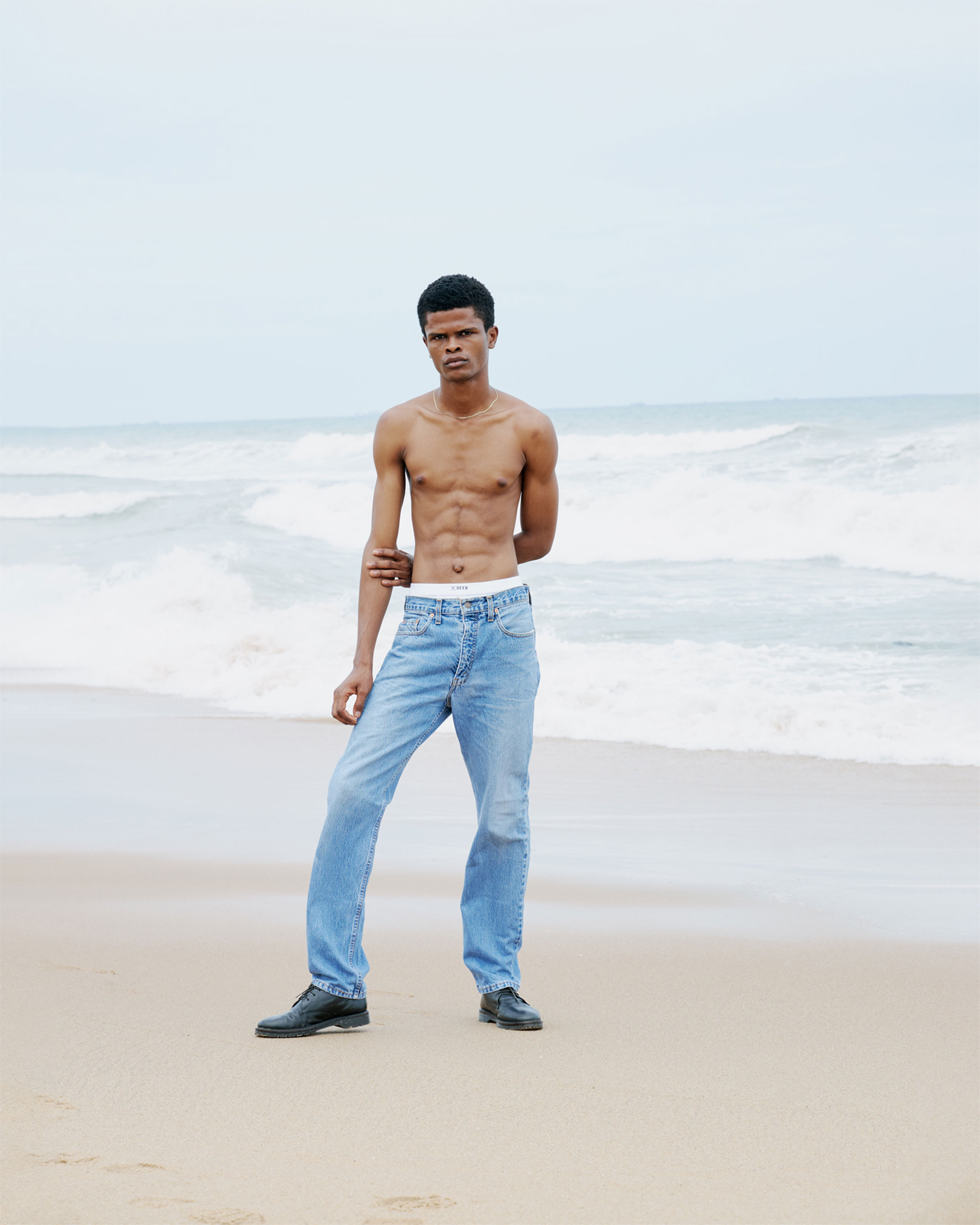 Black man with blue jeans, shirtless, looking straight into the camera, sea and beach in the background, shot by Lisa Marie Asubonteng