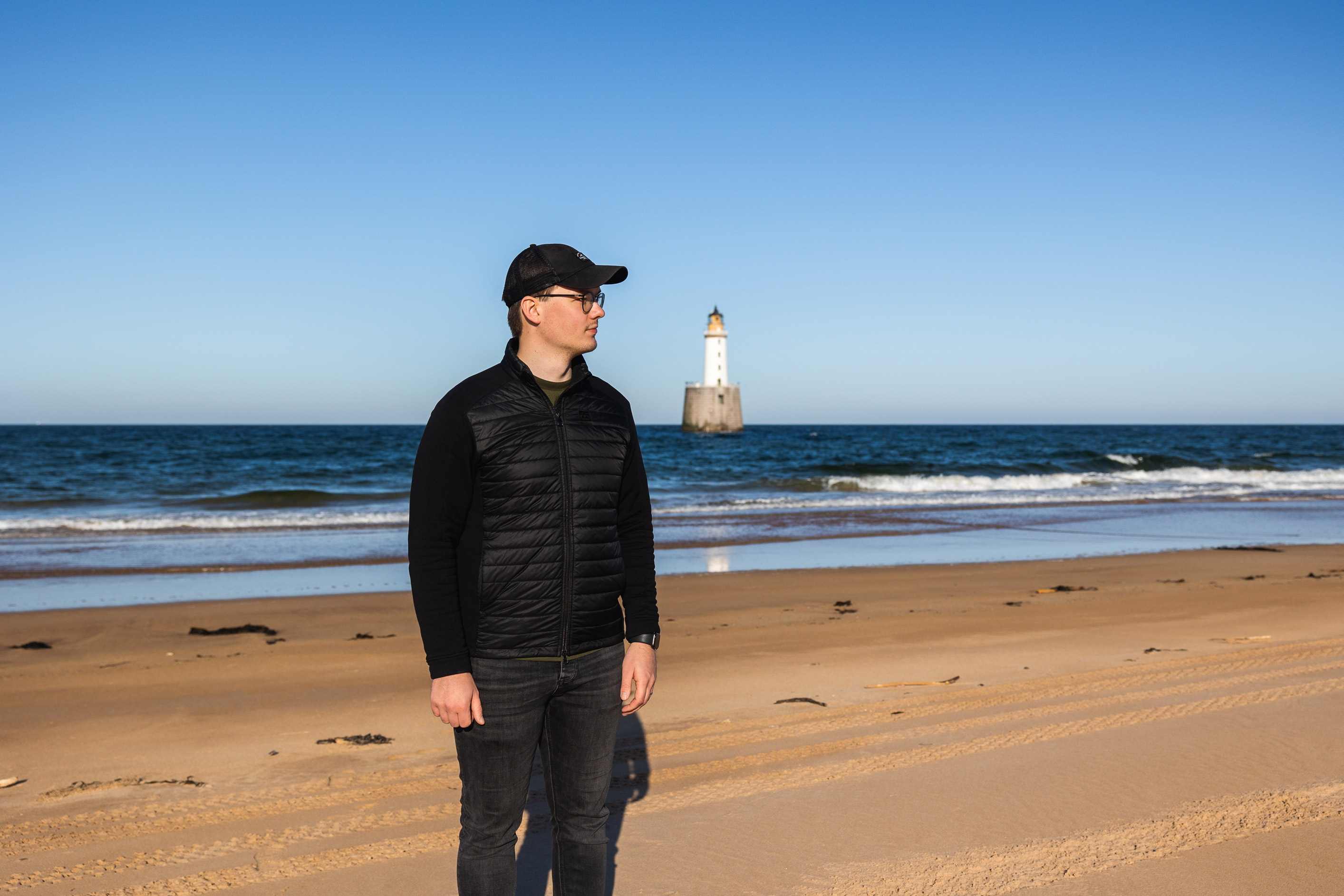 a man wearing a hat standing on a beach with the sea and a lighthouse in the background