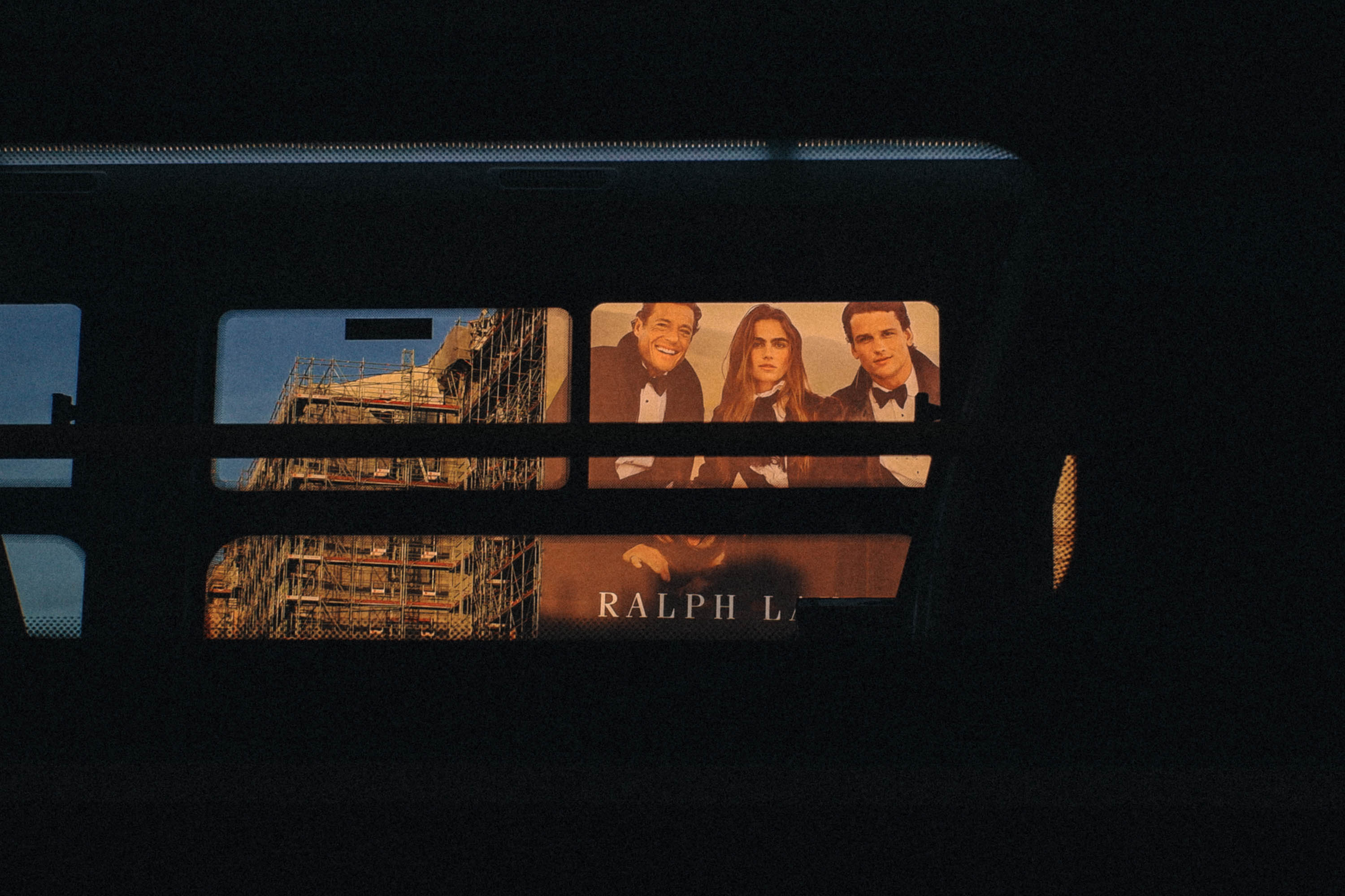 A fashion billboard and scaffolded architecture appearing through the window of a bus, with shadows creating a layered, cinematic composition. Shot by Wesley Verhoeve on a Fujifilm XF10.