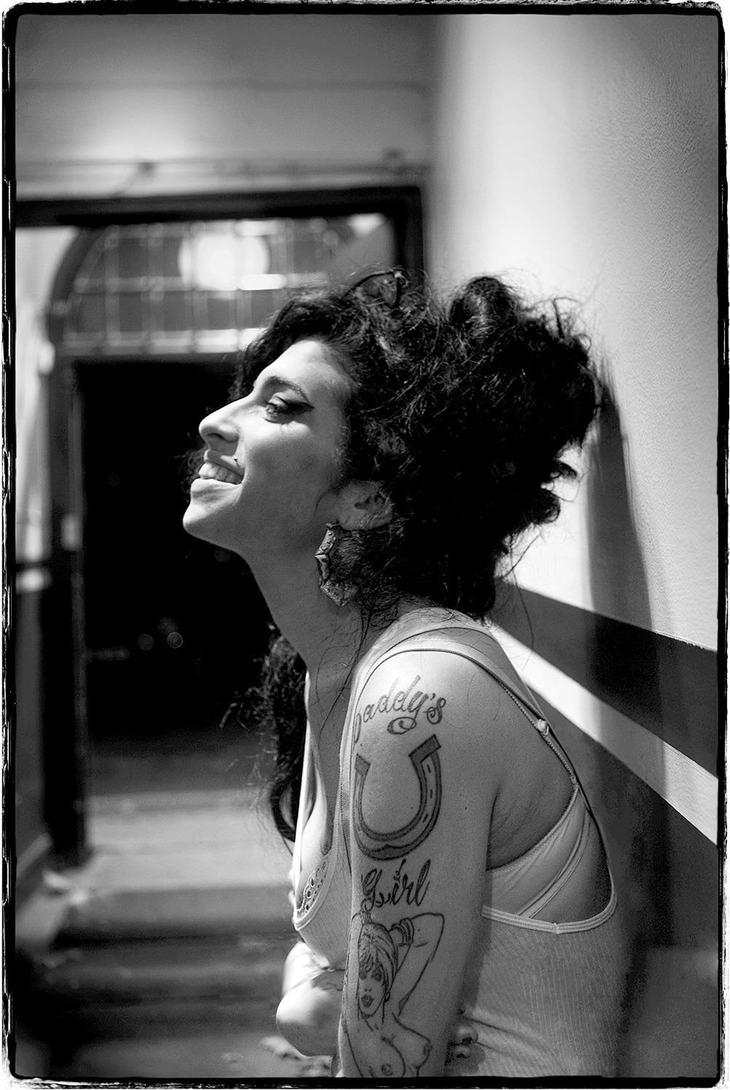 Ein Schwarz-Weiß-Foto von Amy Winehouse, aufgenommen von der Seite, lächelnd, während sie auf einer Bank sitzt, Foto von Jill Furmanovsky